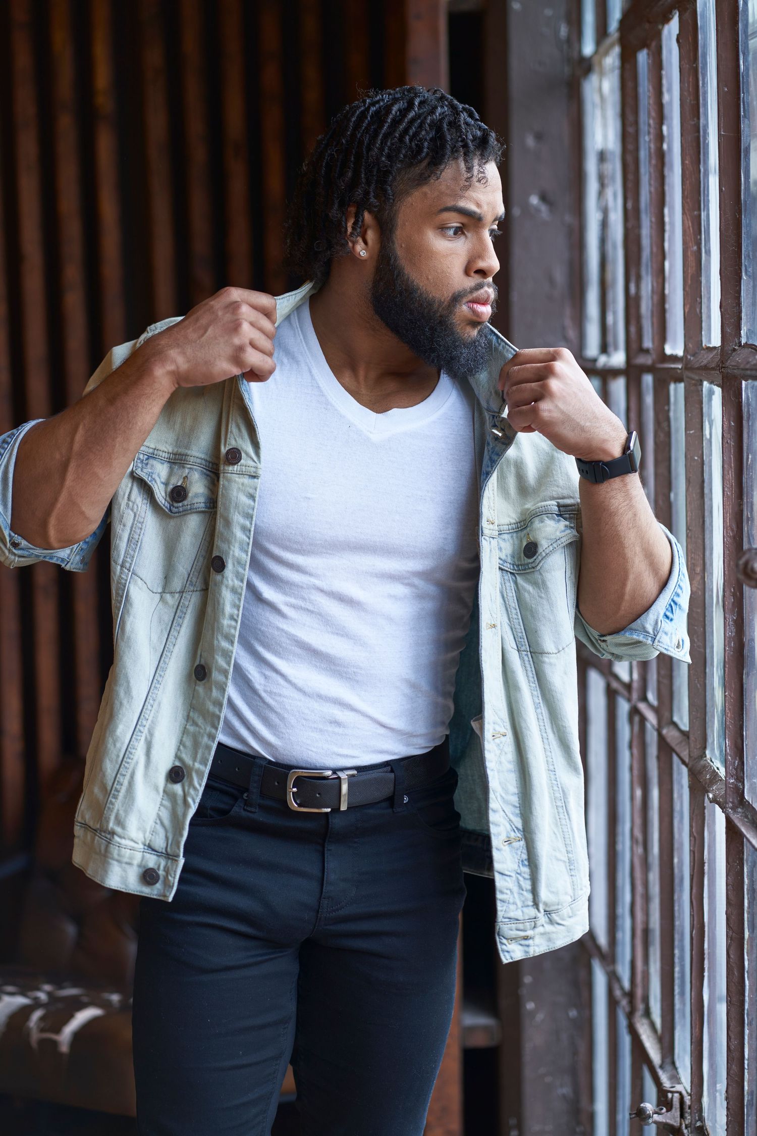 Model in white tank top and light denim jacket poses near window with industrial backdrop.