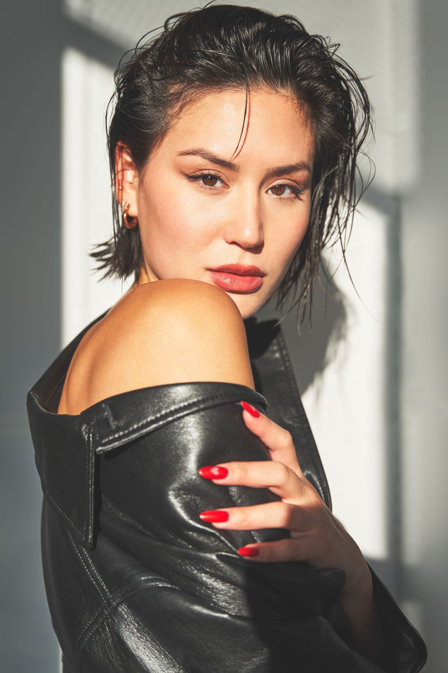 A figure in a black leather jacket poses against a light wall with striking red nail polish and dramatic lighting.