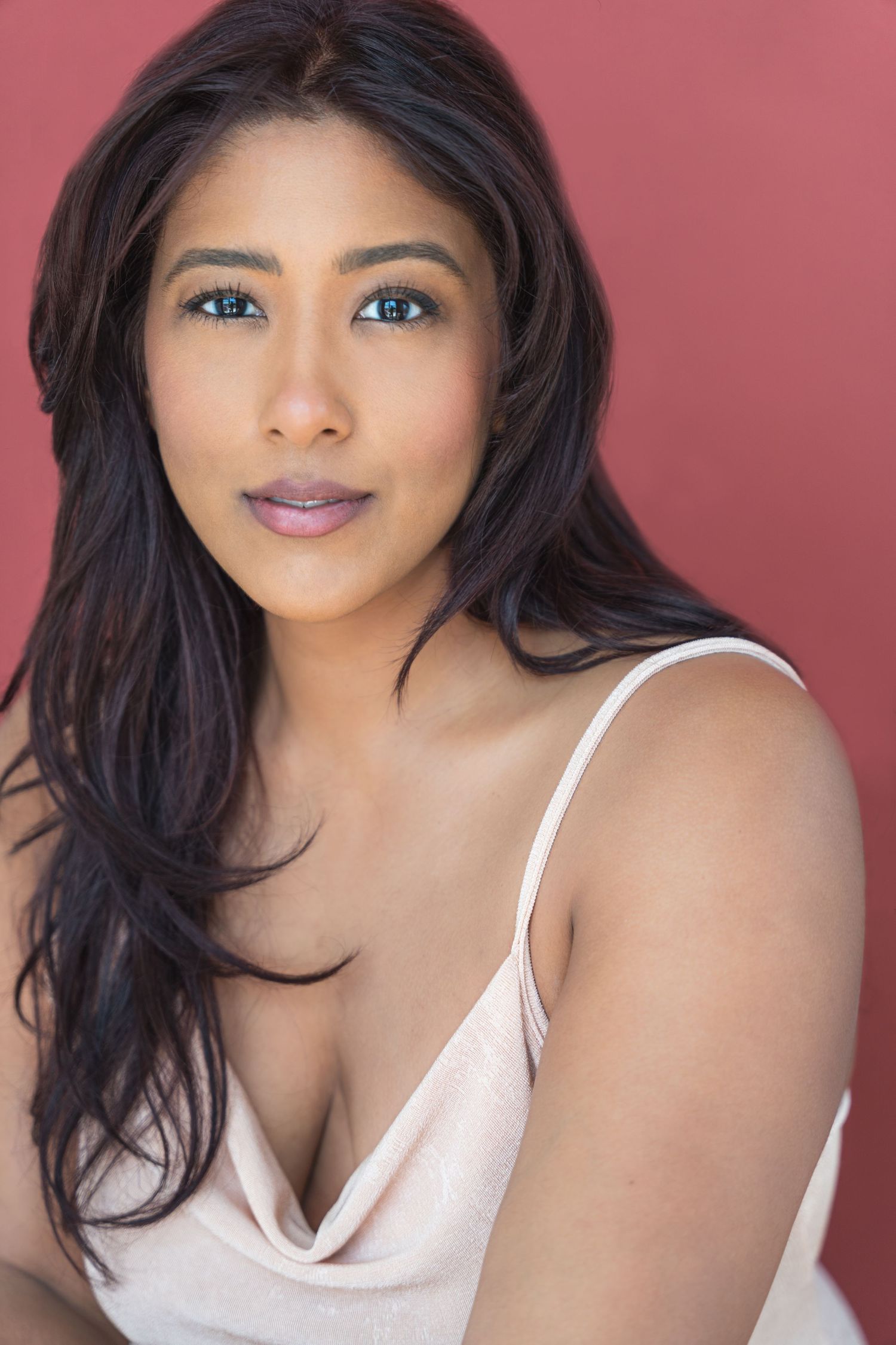 Professional beauty portrait of woman with long dark hair wearing white top against pink background.
