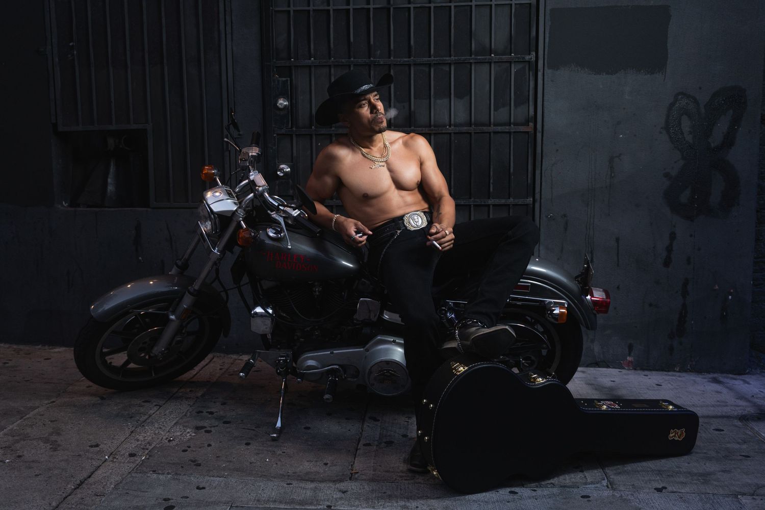 A shirtless motorcyclist sits on a parked motorcycle next to a guitar case in a dark urban setting.