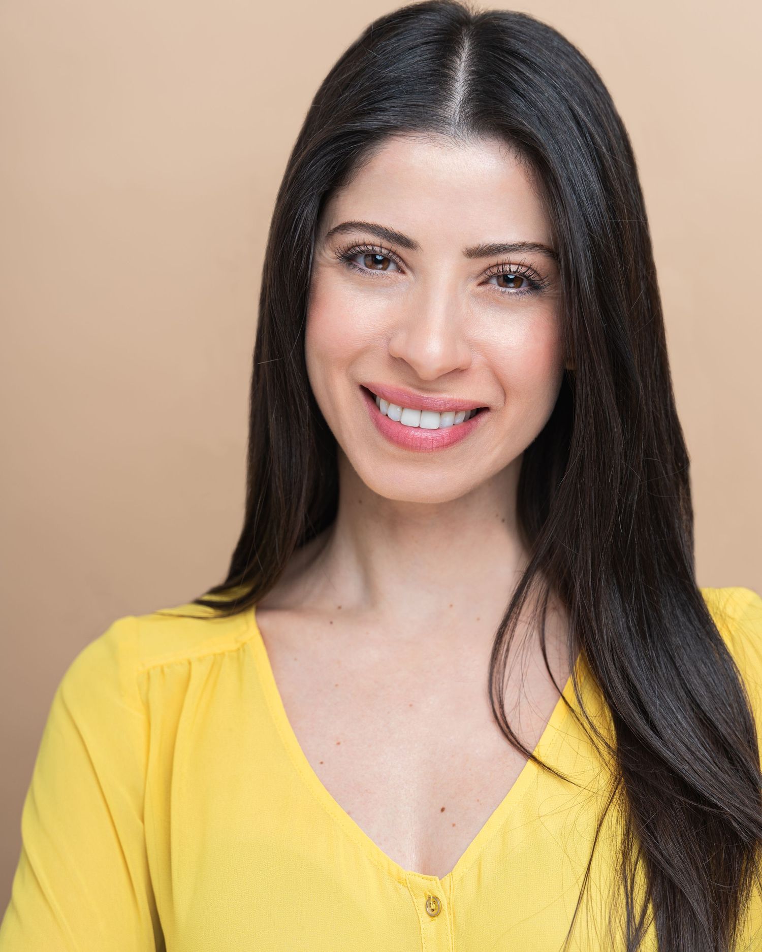 Portrait of someone with long dark hair and a bright yellow v-neck top smiling warmly against a neutral background.