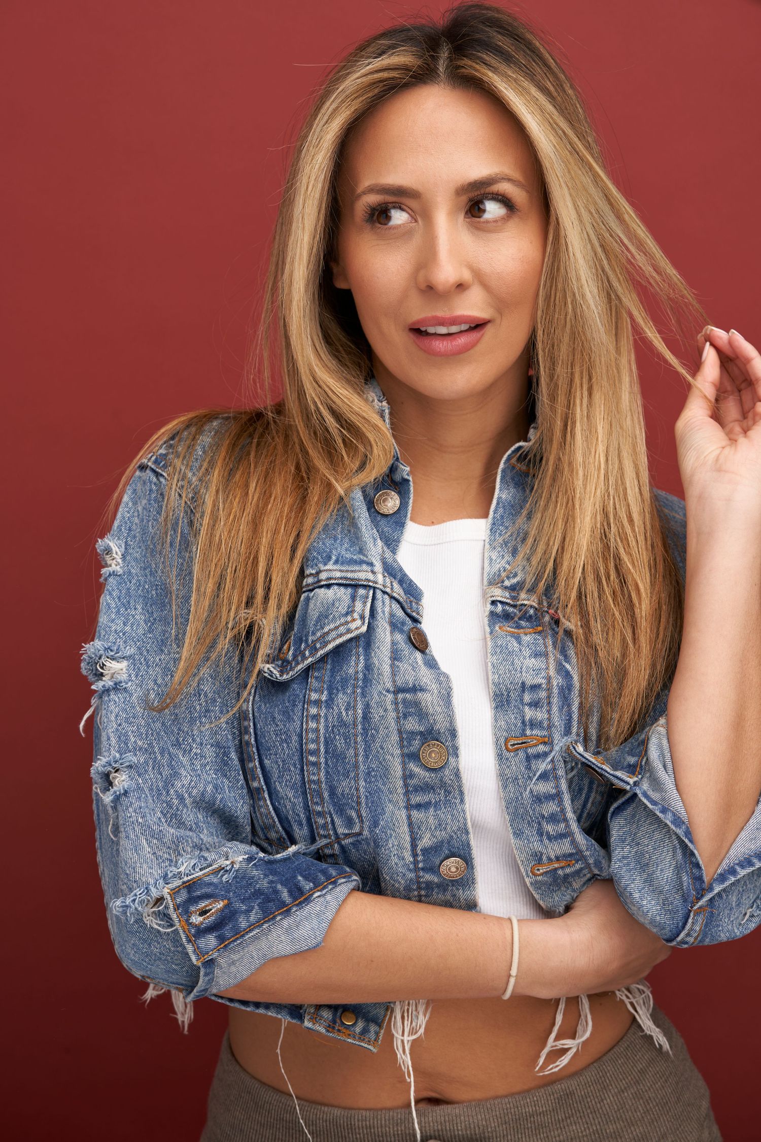 Young blonde woman wearing distressed denim jacket and white top poses playfully against burgundy background.