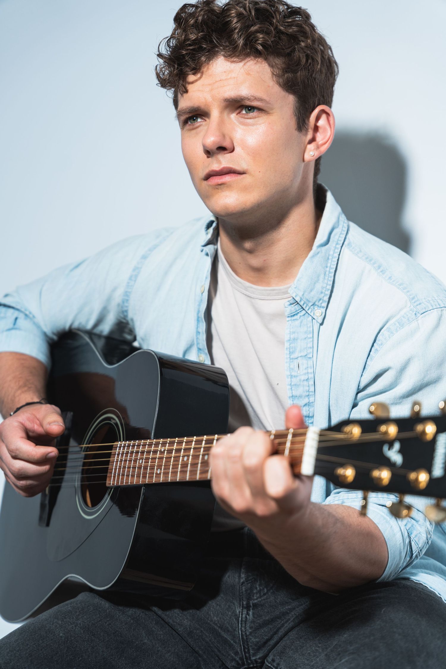 Musician in light denim shirt playing black acoustic guitar against white background.