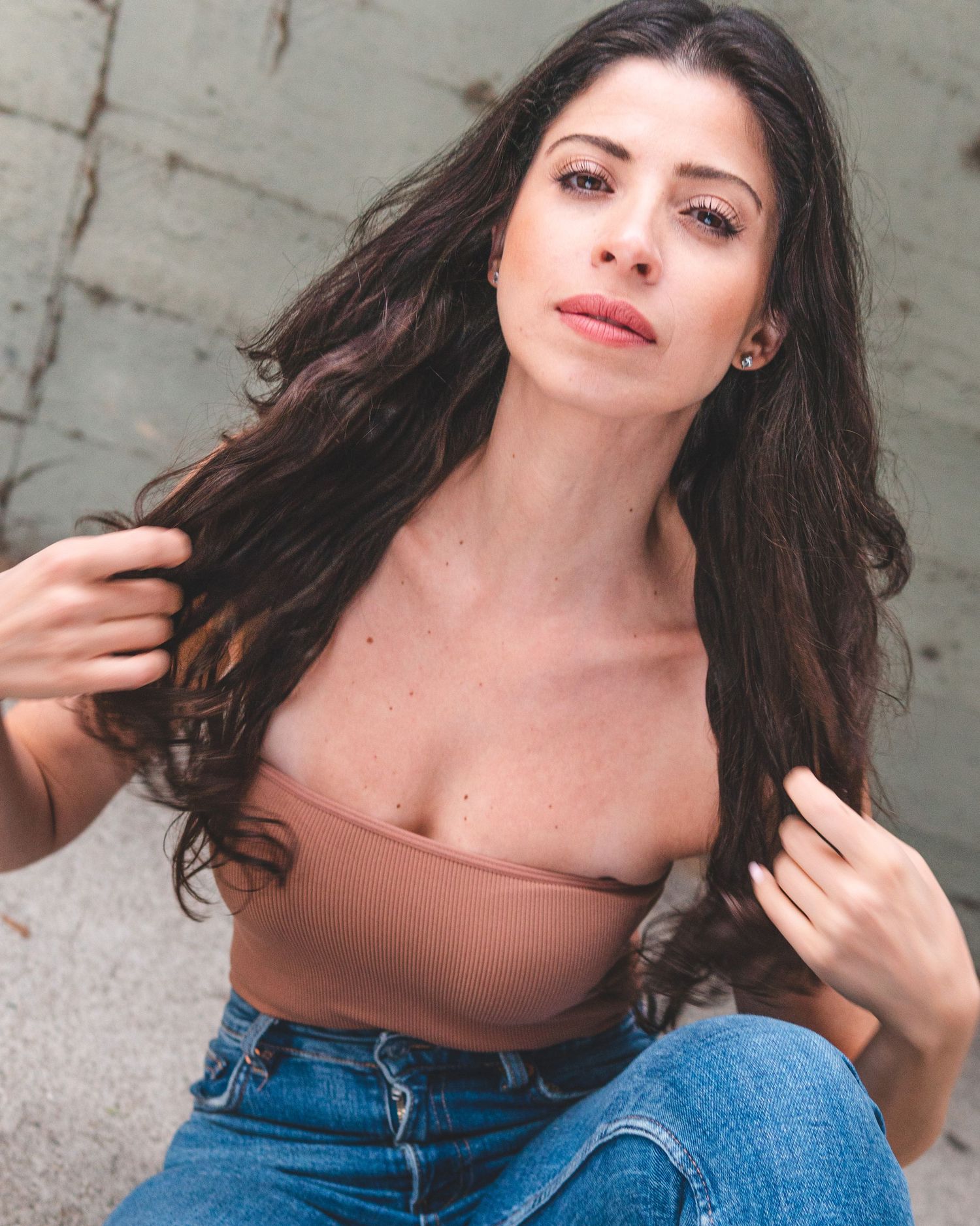 A stunning portrait of a woman with long dark hair wearing a brown crop top and jeans against a gray background.