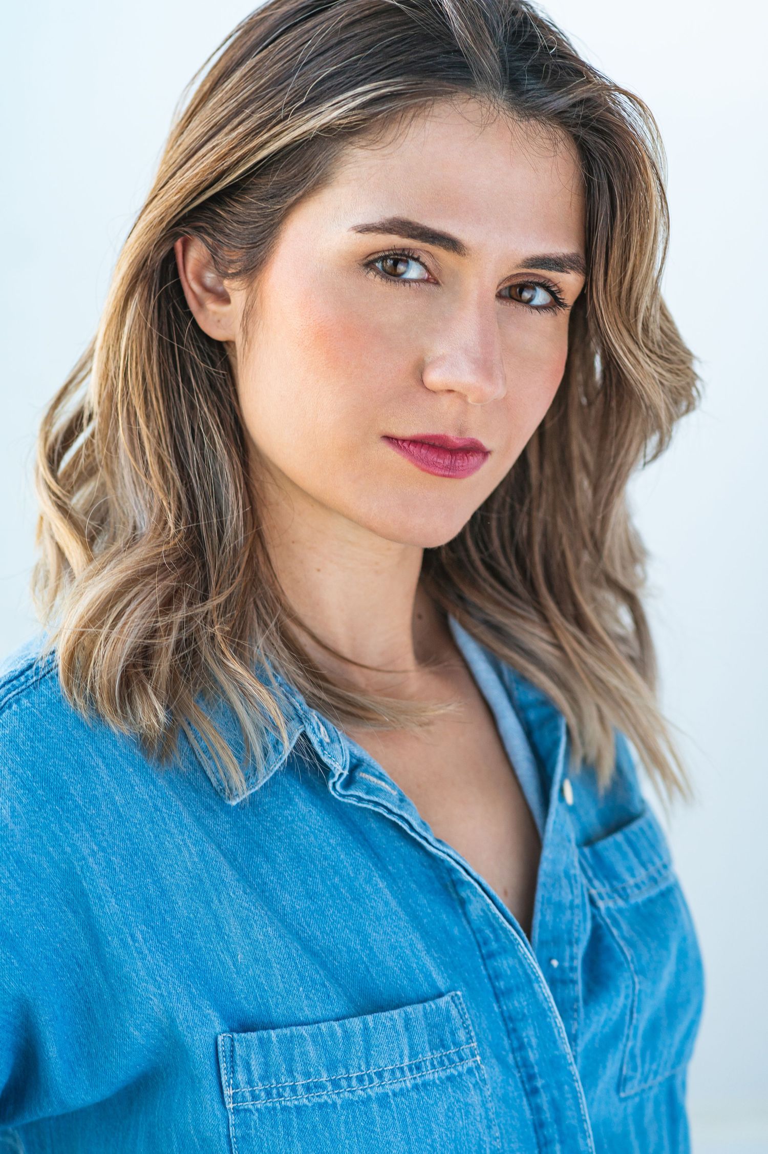 Portrait of someone wearing a casual denim shirt against white background with wavy brown hair and subtle makeup.