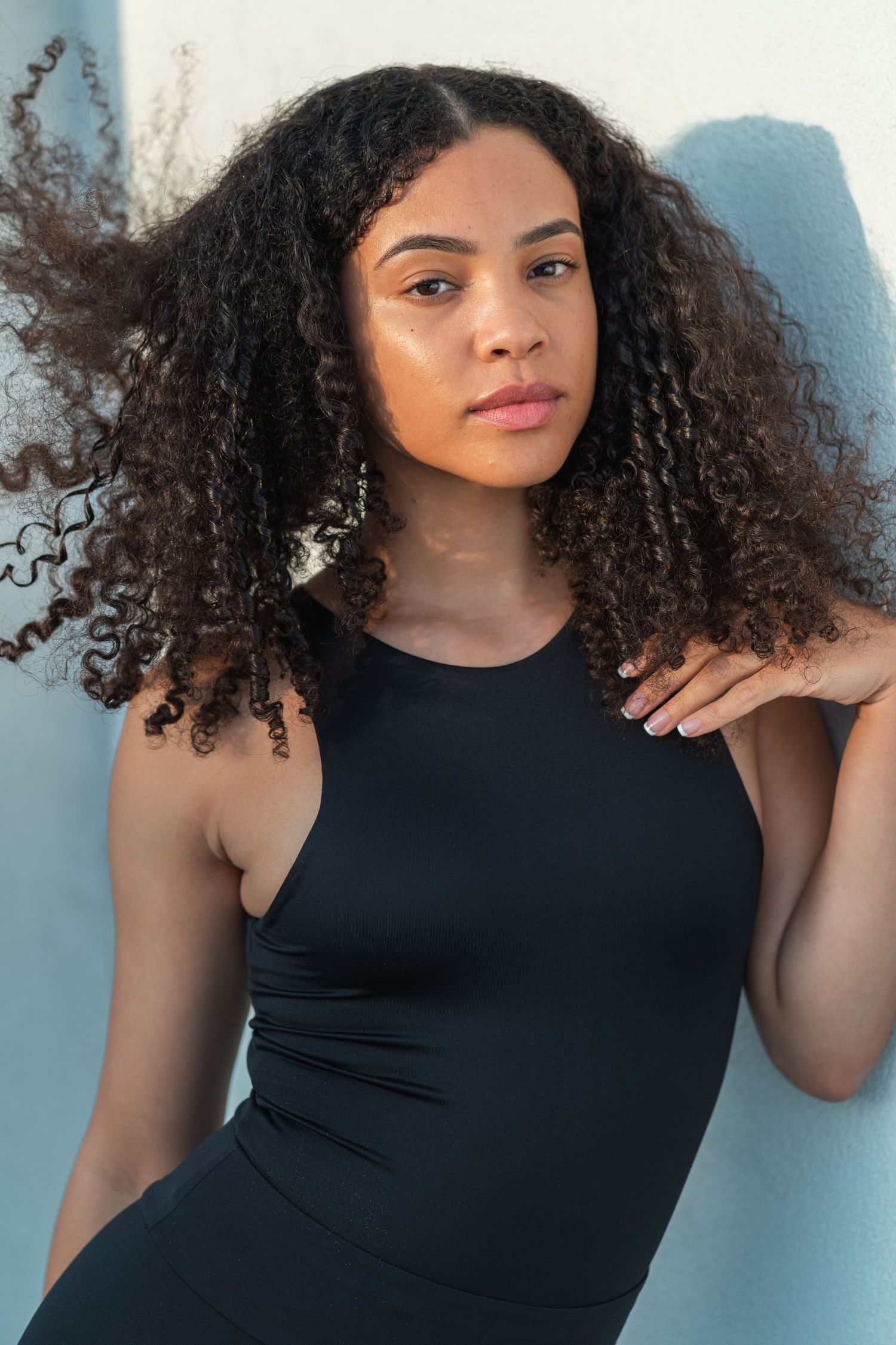 Model in black tank top with natural curly hair poses against light blue background.