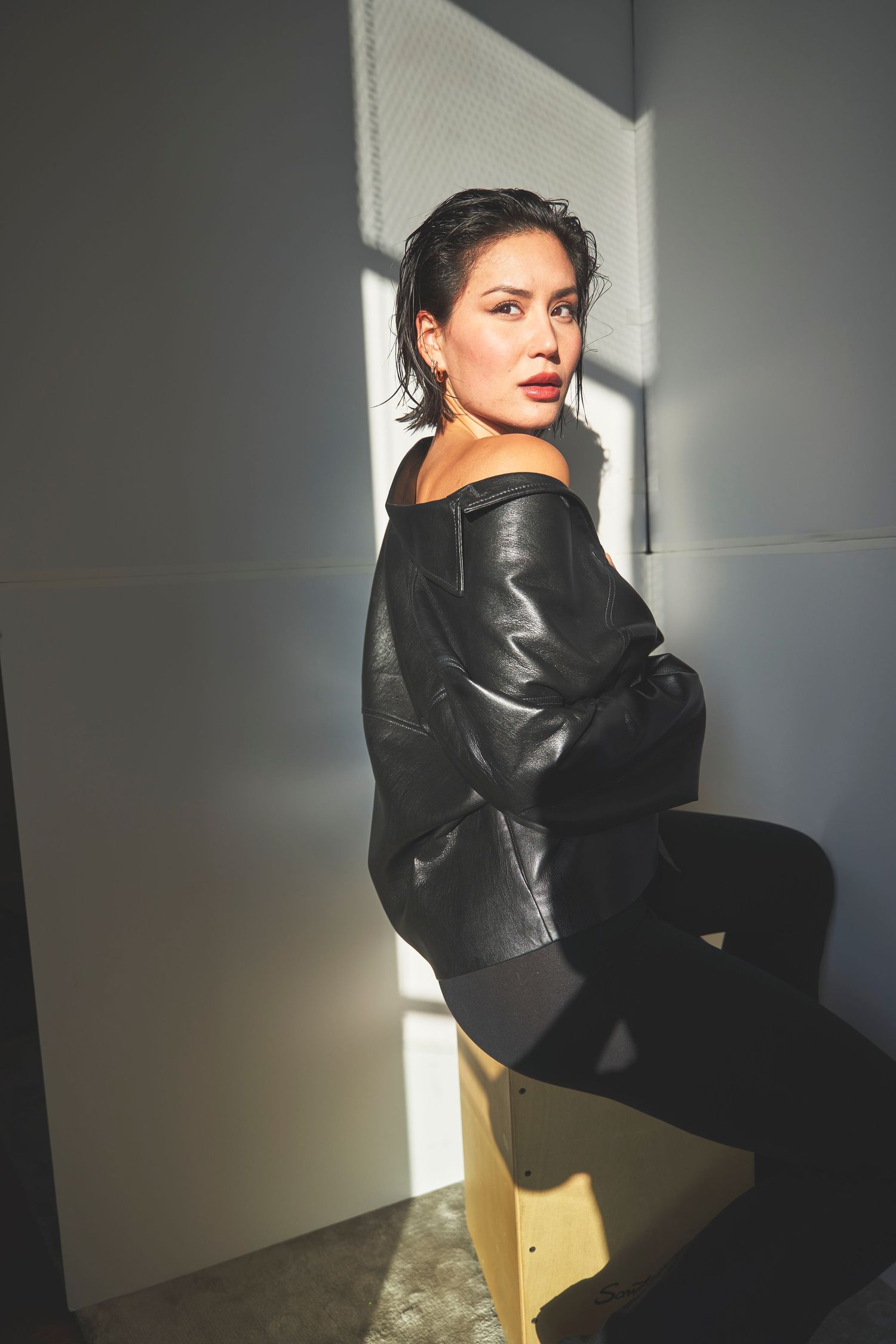Fashion portrait of model in black leather jacket sitting in dramatic lighting against white wall.
