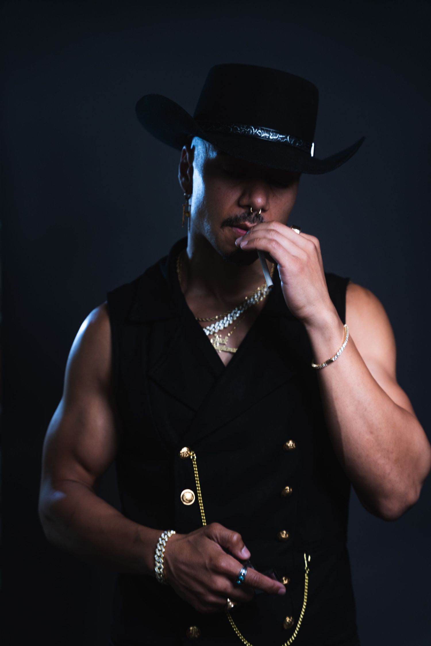Dramatic studio portrait with moody lighting shows muscular figure in black hat holding a metal chain against dark background.