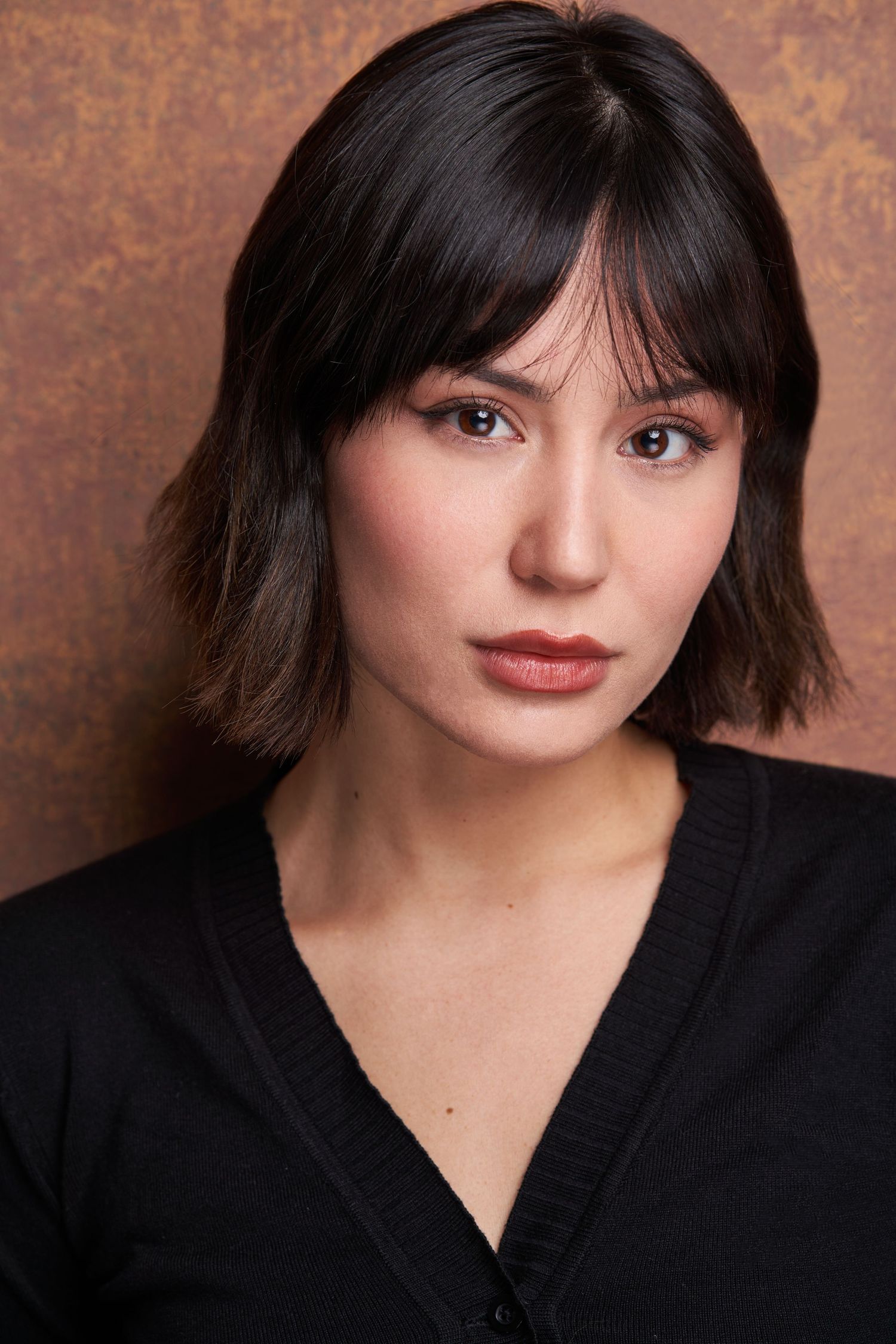 Portrait against a warm brown textured backdrop featuring dark hair in a bob cut and black v-neck sweater.