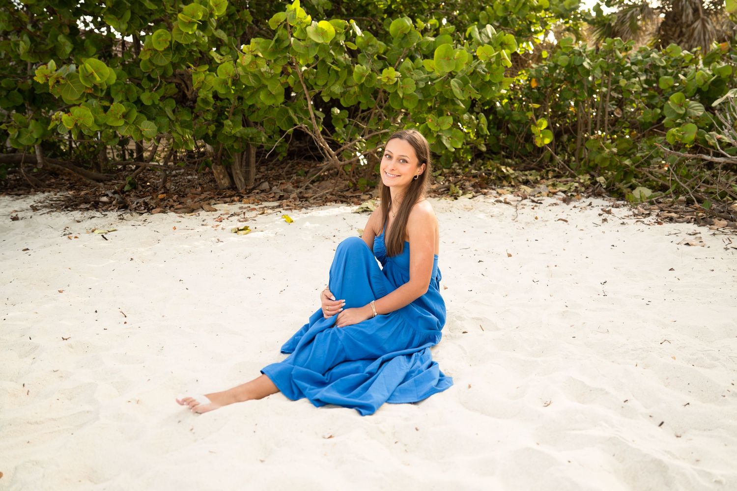 Woman in blue dress sitting on white sand beach with lush green tropical trees in background. Naples, Florida.