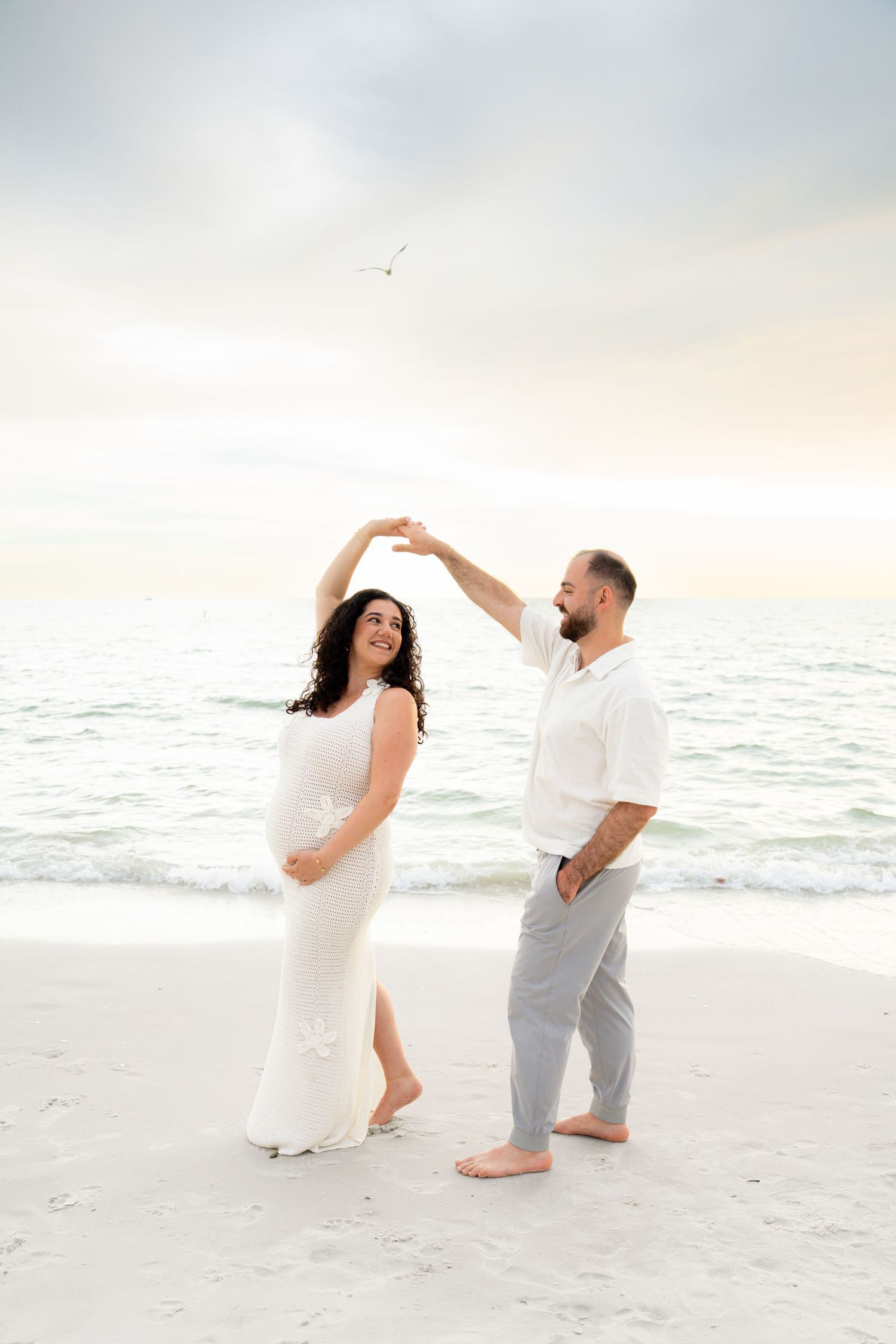 Pregnant couple dancing on a beach at sunset, woman in white dress, man in white shirt and gray pants. Naples, Florida.