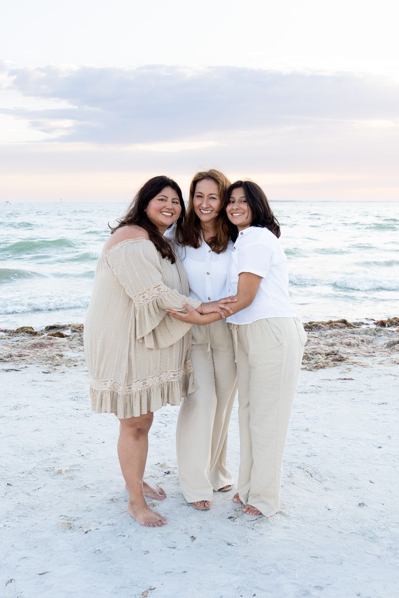 Three women holding hands and smiling together on a beach during a soft sunset. Fort Myers Beach, Florida.