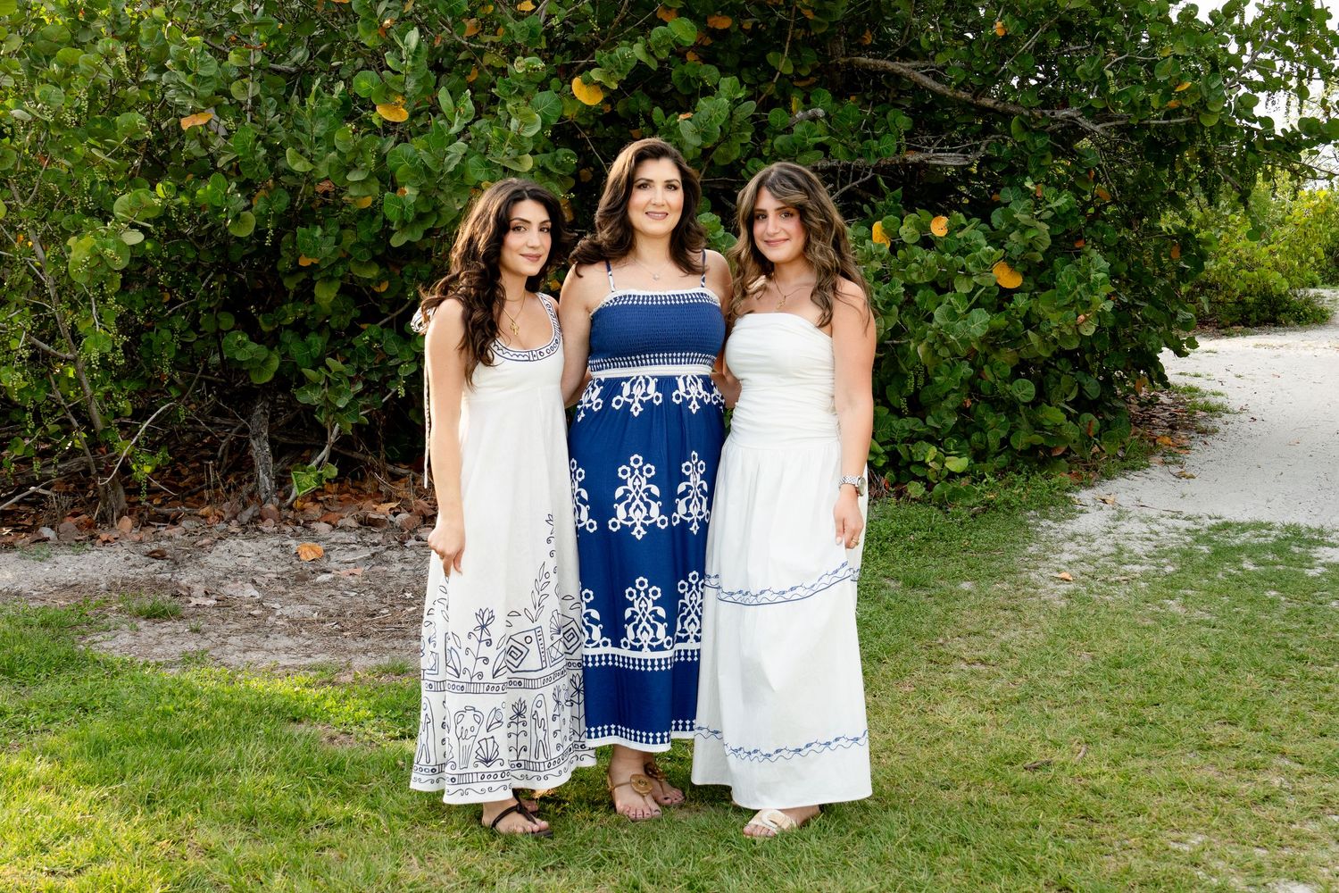 Three women in white and blue maxi dresses posing outdoors near lush green tropical trees.