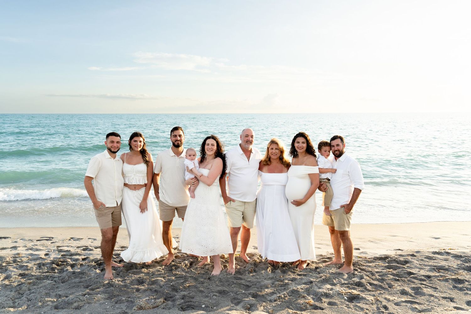Large family in white outfits posing together on a sunny beach with ocean waves in the background.