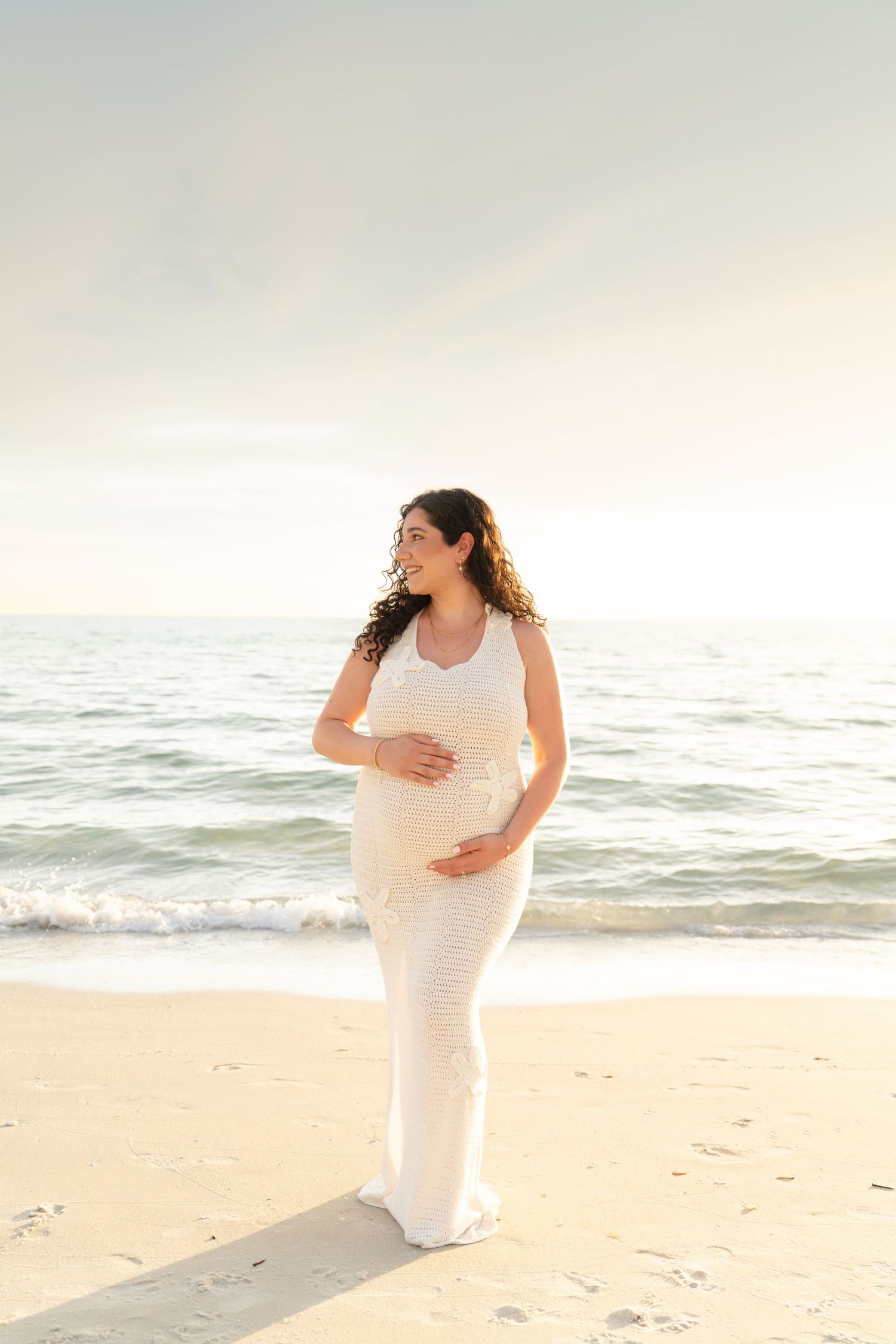 Pregnant woman in white dress cradling baby bump on sandy beach at golden hour with ocean waves behind her. Naples, Florida.