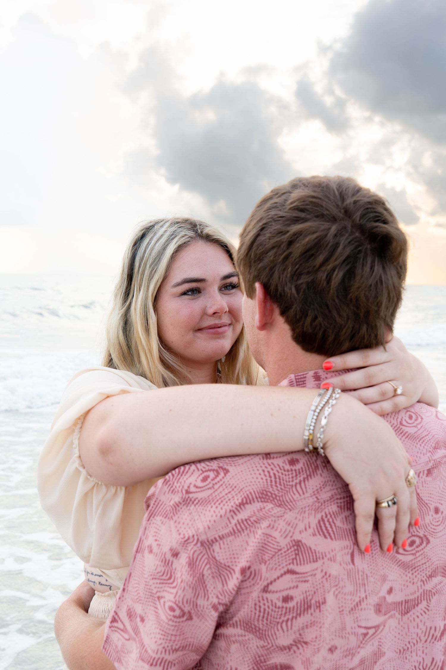 Couple embracing on a beach at sunset, woman with blonde hair smiling, wearing a ring and bracelet. Proposal in Bonita Springs, Florida.