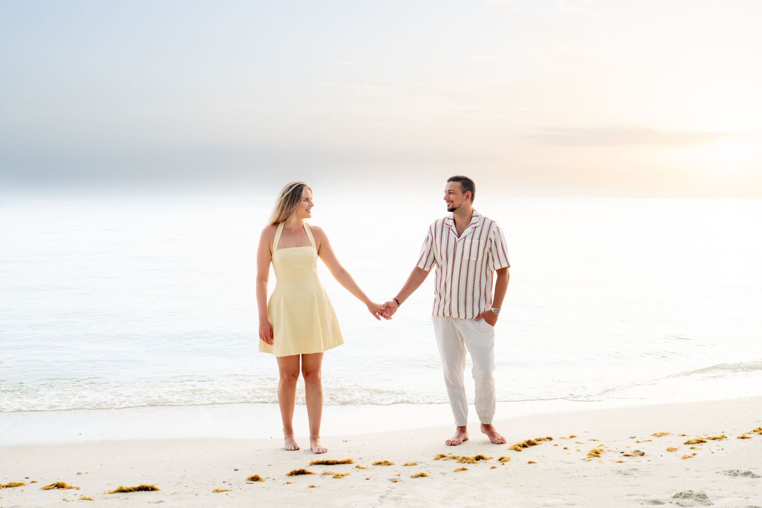 Couple holding hands on a sandy beach at sunset, woman in yellow dress, man in striped shirt, ocean in background. Naples, Florida.