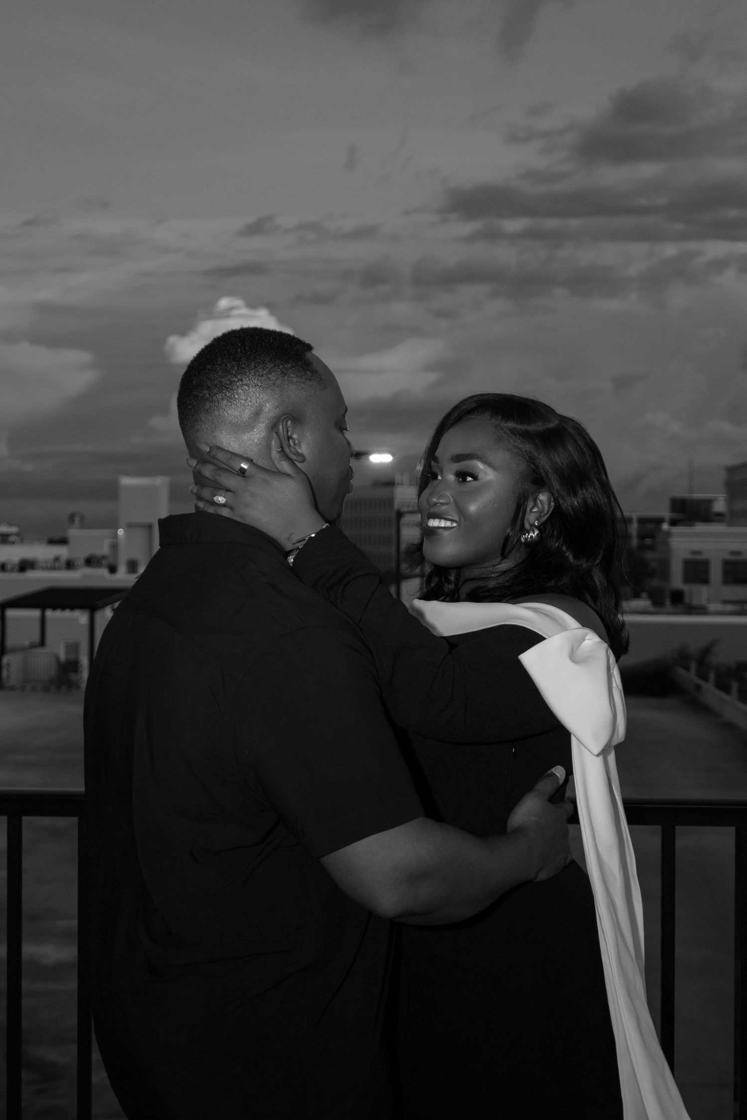 Black and white photo of a couple embracing romantically on a rooftop at dusk.