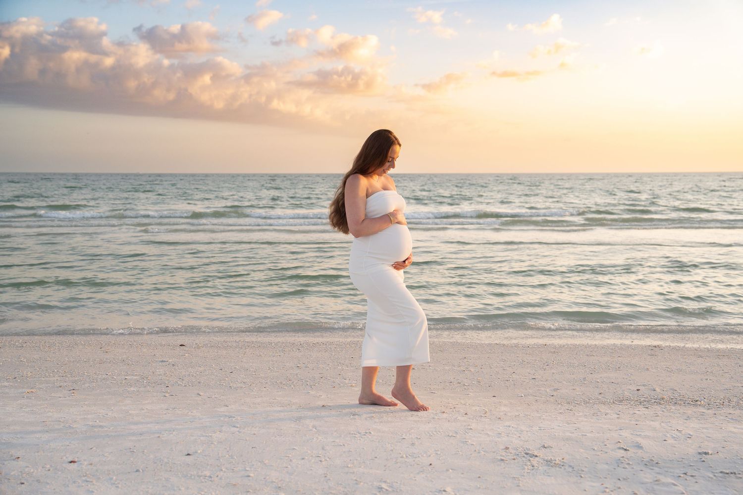 Pregnant woman in white dress cradling baby bump on white sand beach at golden sunset.