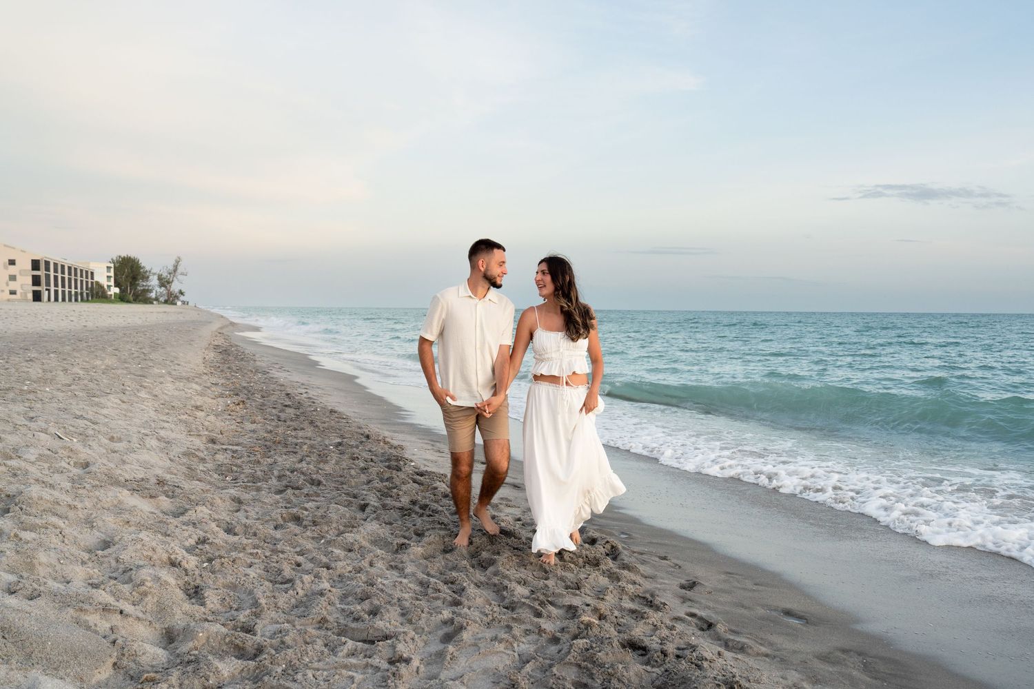 Happy couple in white outfits walking hand in hand along sandy beach shoreline at sunset.