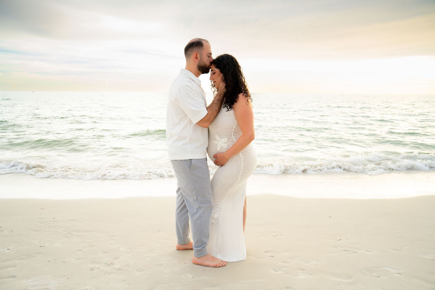 Couple embracing on a sandy beach at sunset, woman in white maternity dress, man in white shirt and gray pants.