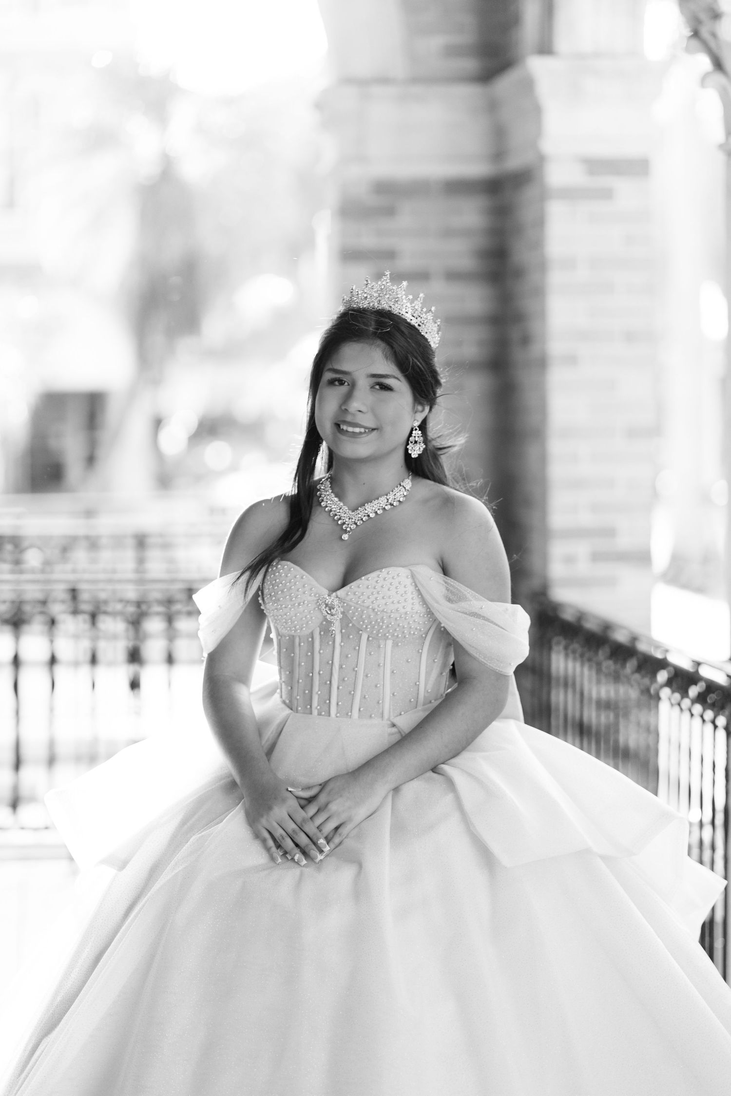 Black and white portrait of a young woman in a quinceañera gown and tiara on a balcony.