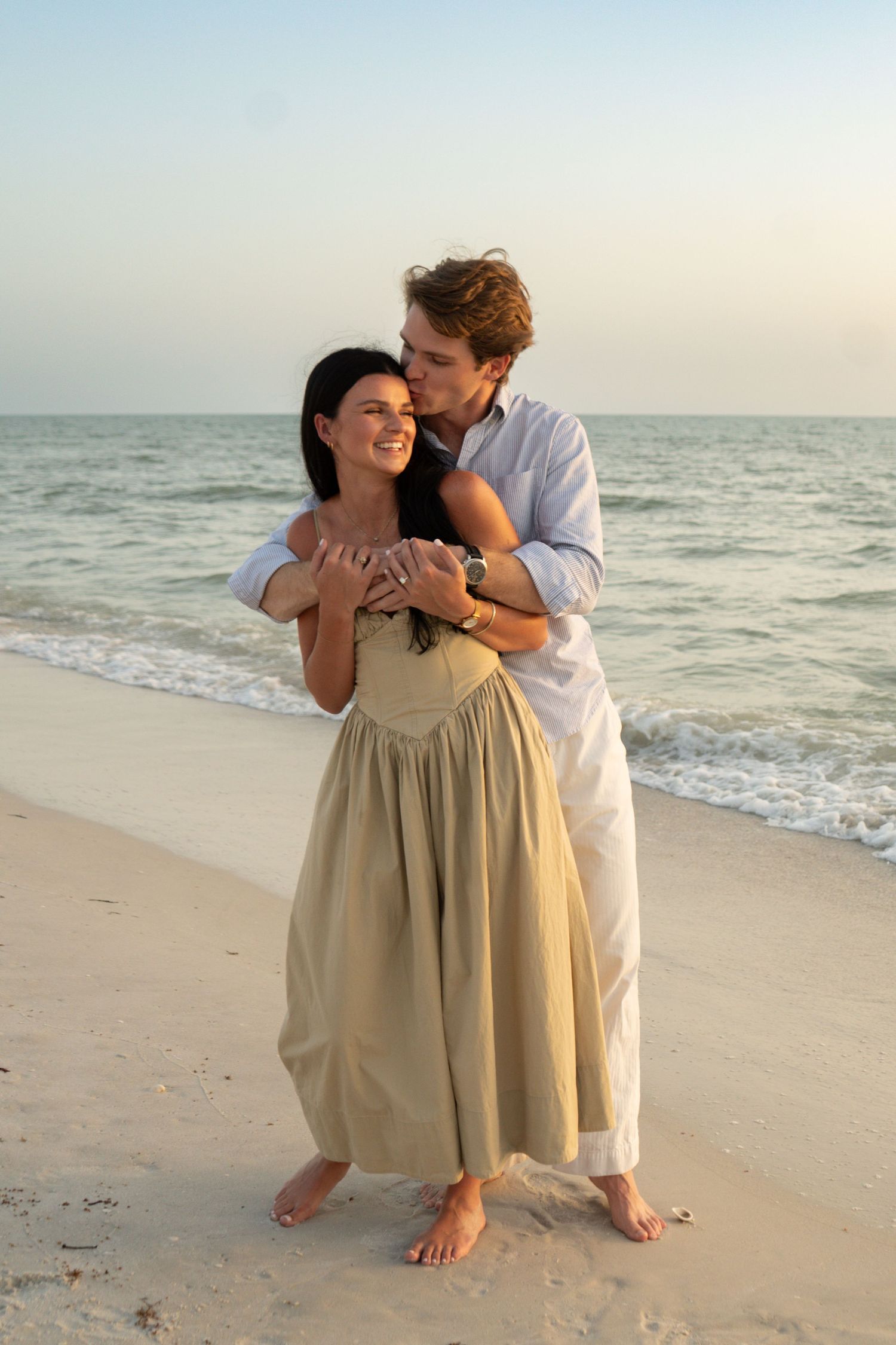 Romantic couple embracing on a sandy beach at sunset, woman in elegant dress, man kissing her cheek near ocean waves.