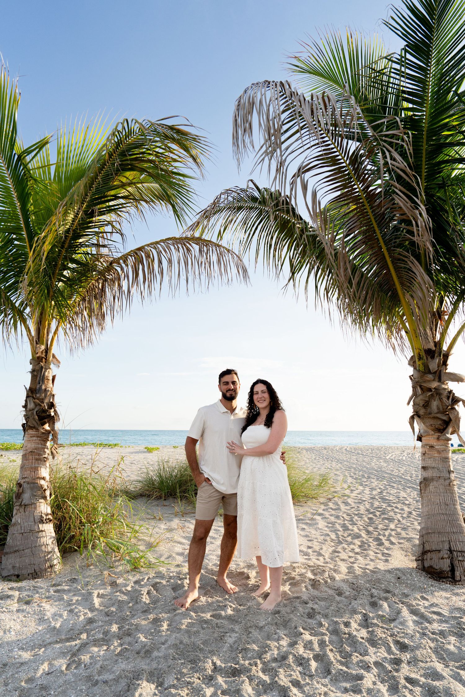 Couple standing between two palm trees on a sandy beach with clear blue skies and ocean in the background. Marco Island, Florida.