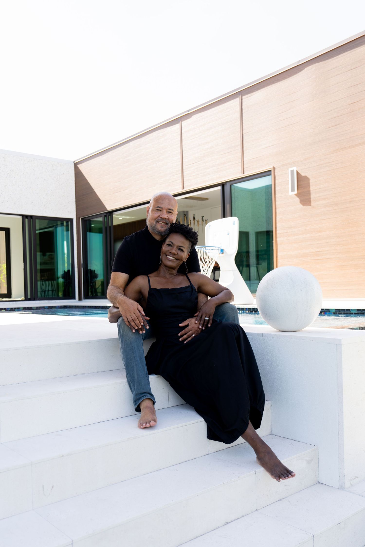 A smiling couple in black outfits sits on white steps outside a modern home with a pool. Marco Island, Florida.