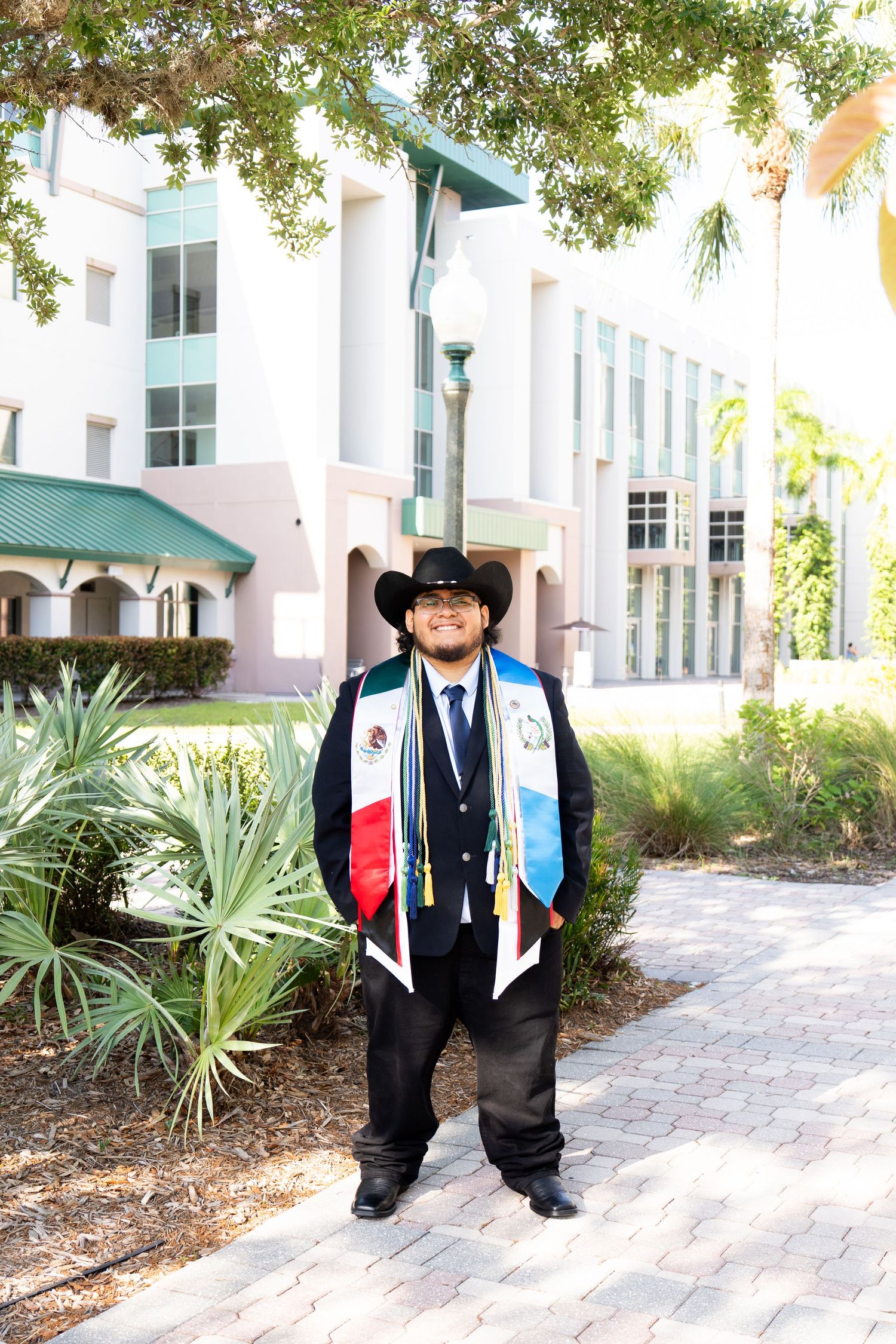 Graduate in black suit, cowboy hat, and colorful stoles posing on a sunny college campus. Florida Gulf Coast University Graduation Photos.