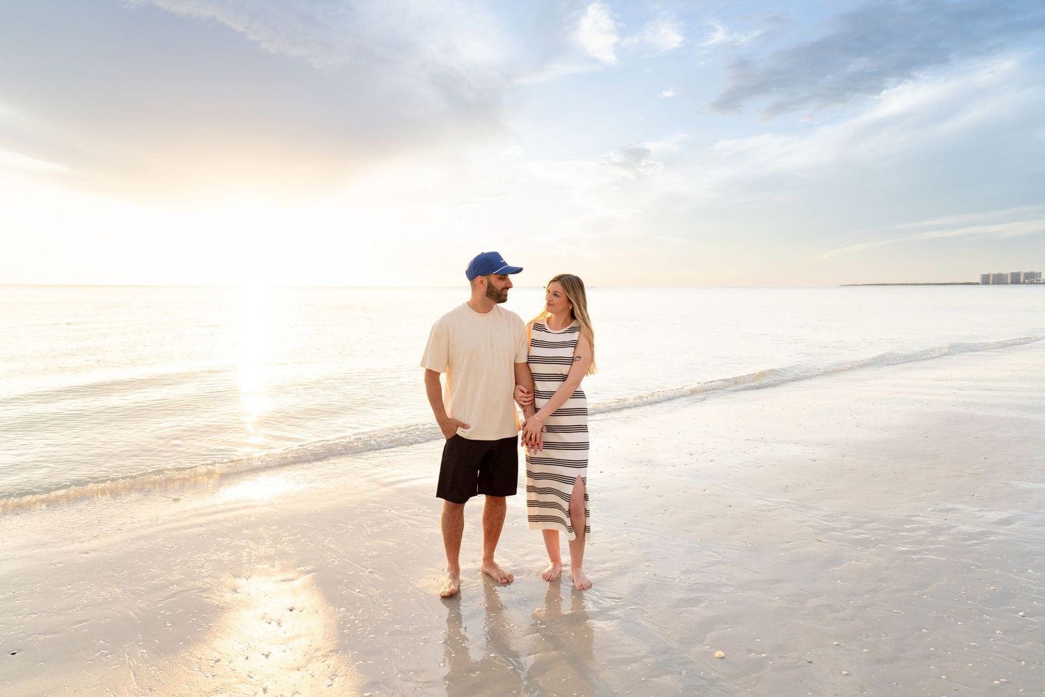 Couple holding hands on a sunny beach at sunset, man in blue cap and woman in striped dress, waves gently lapping shore.