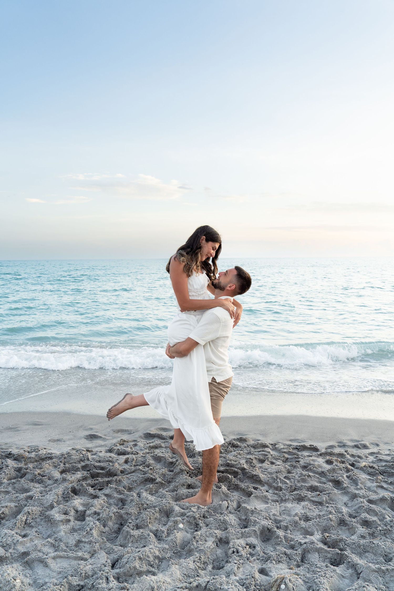 Romantic couple at beach as man lifts woman in white dress near turquoise ocean waves at sunset.