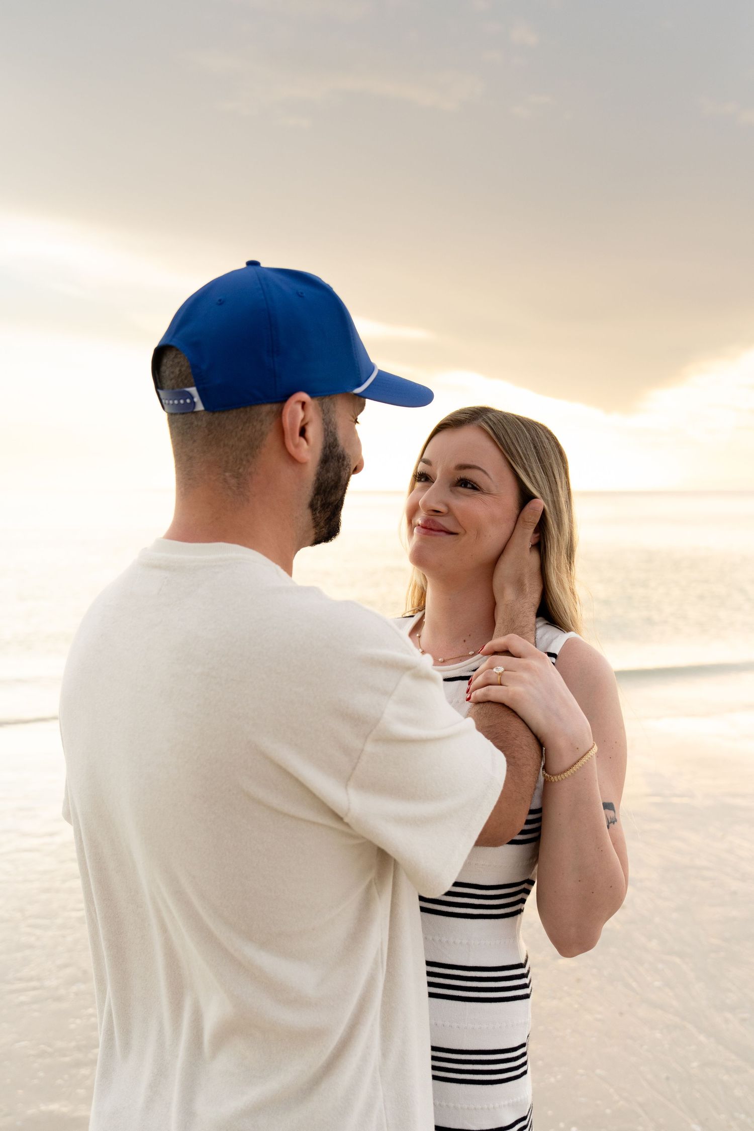 Couple sharing a romantic moment on the beach at sunset, woman in striped dress smiling at bearded man in blue cap. Bonita Springs, Florida.