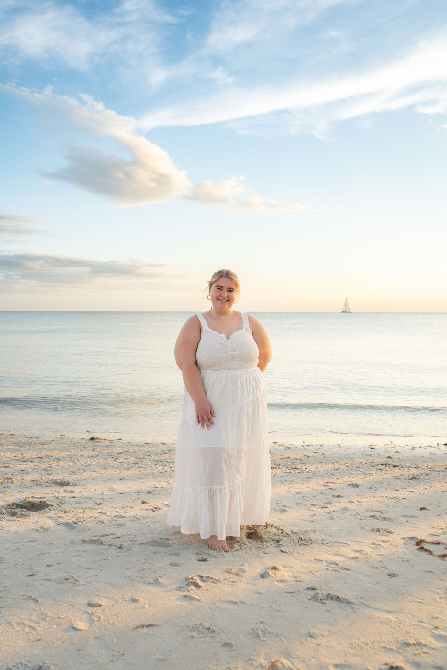 Woman in white maxi dress standing on sandy beach at sunset with calm ocean and sailboat in background. Naples, Florida.