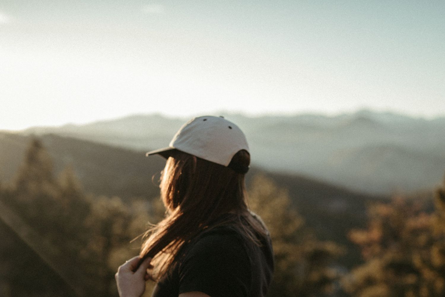 A person in a baseball cap looks out over a scenic mountain landscape during golden hour.