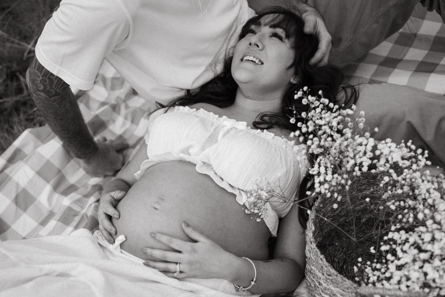 A pregnant woman in a white lace top lies on a checkered blanket with baby's breath flowers beside her.