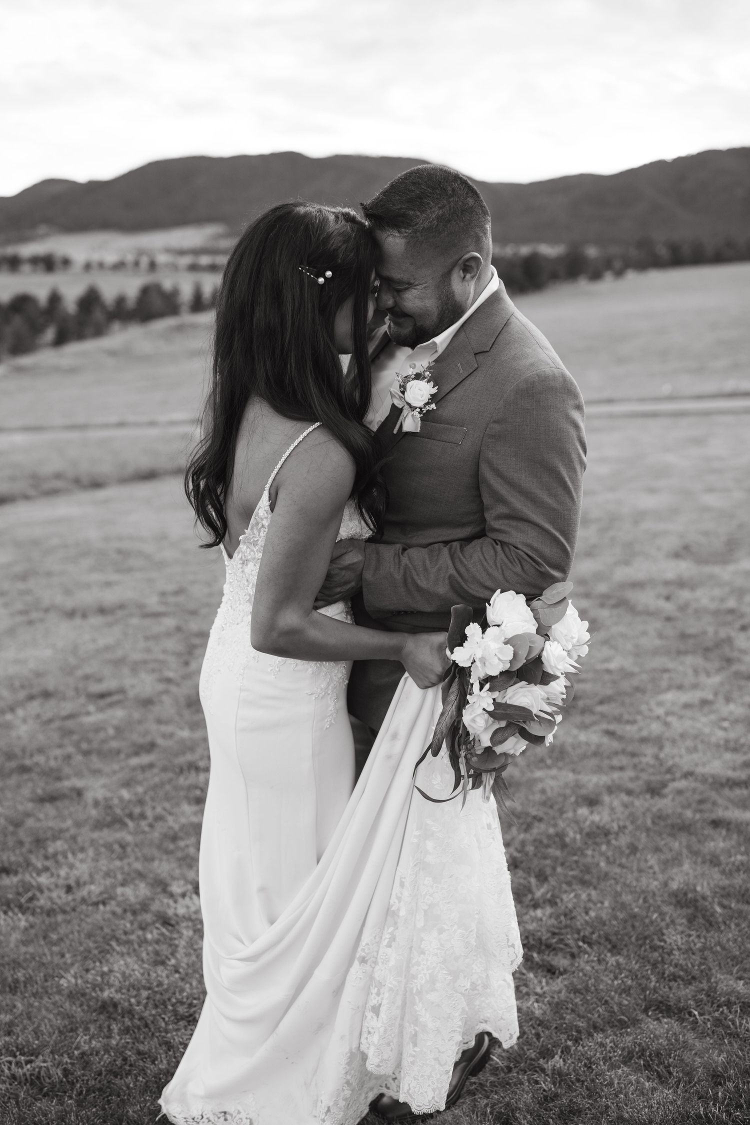 Black and white portrait of newlyweds embracing in a mountain field with white floral bouquet.
