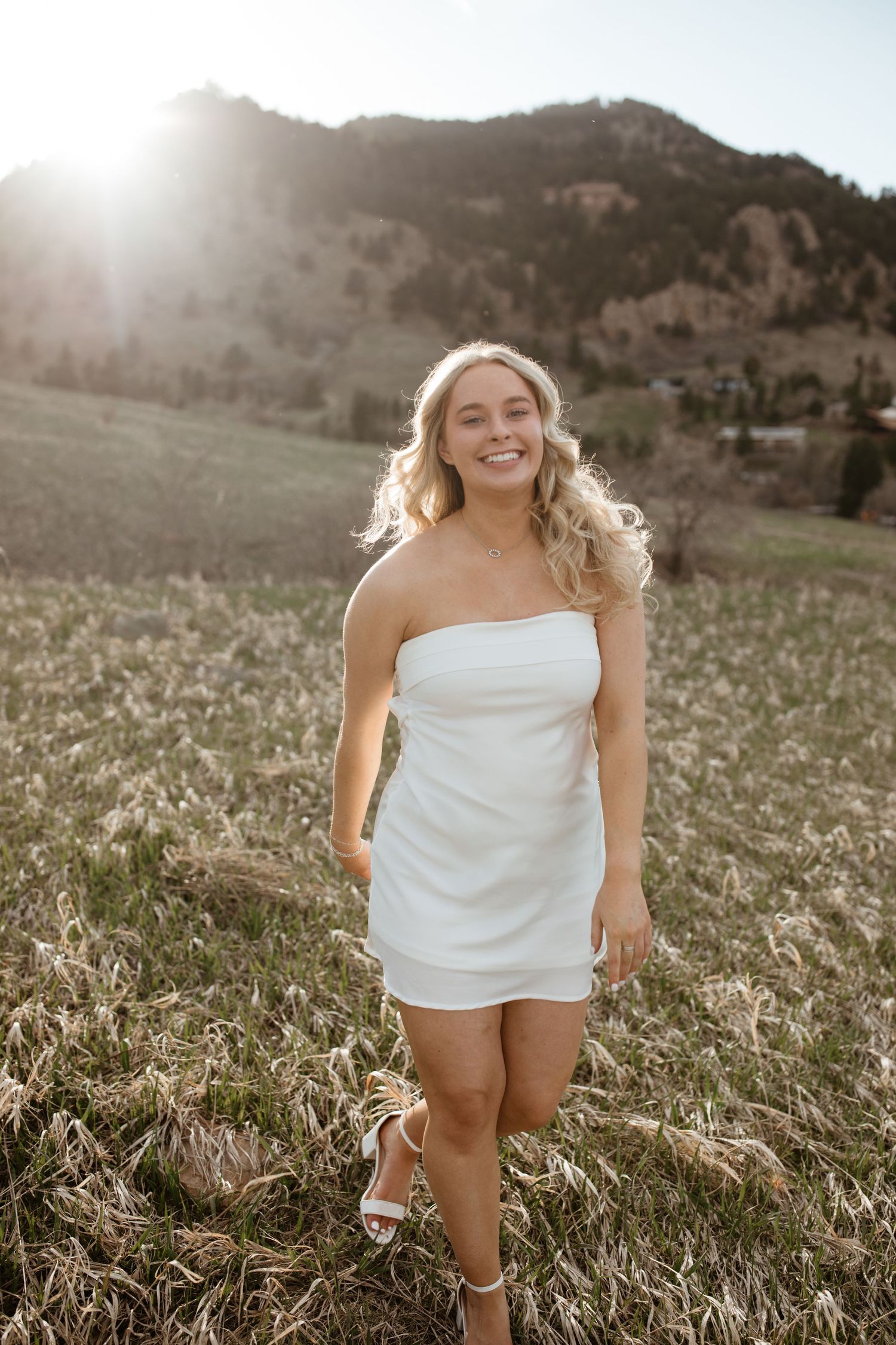 Woman walking through mountain meadow wearing short white dress at sunset.
