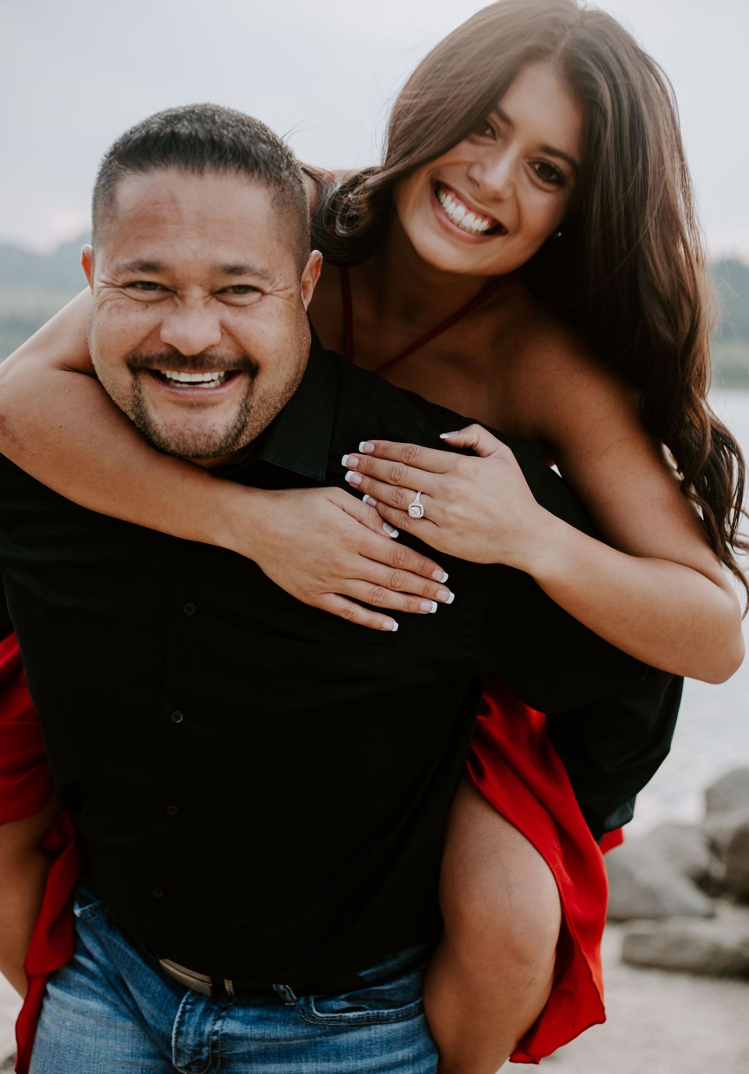 Joyful piggyback engagement photo showing off ring in red dress against nature backdrop.