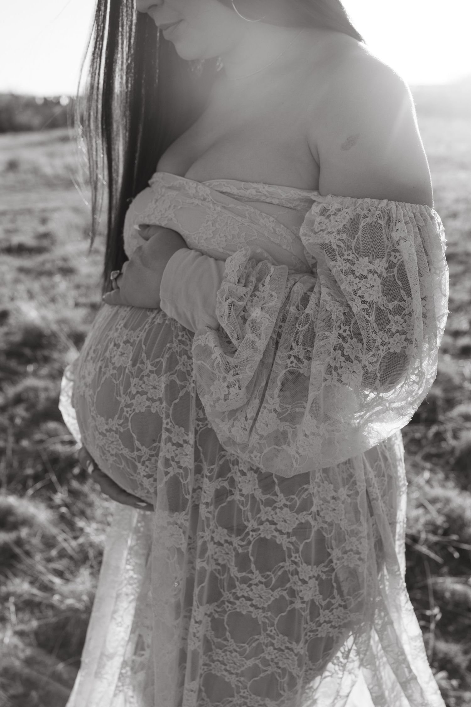Black and white maternity photo of pregnant woman in lace off-shoulder gown cradling baby bump in sunlit field.