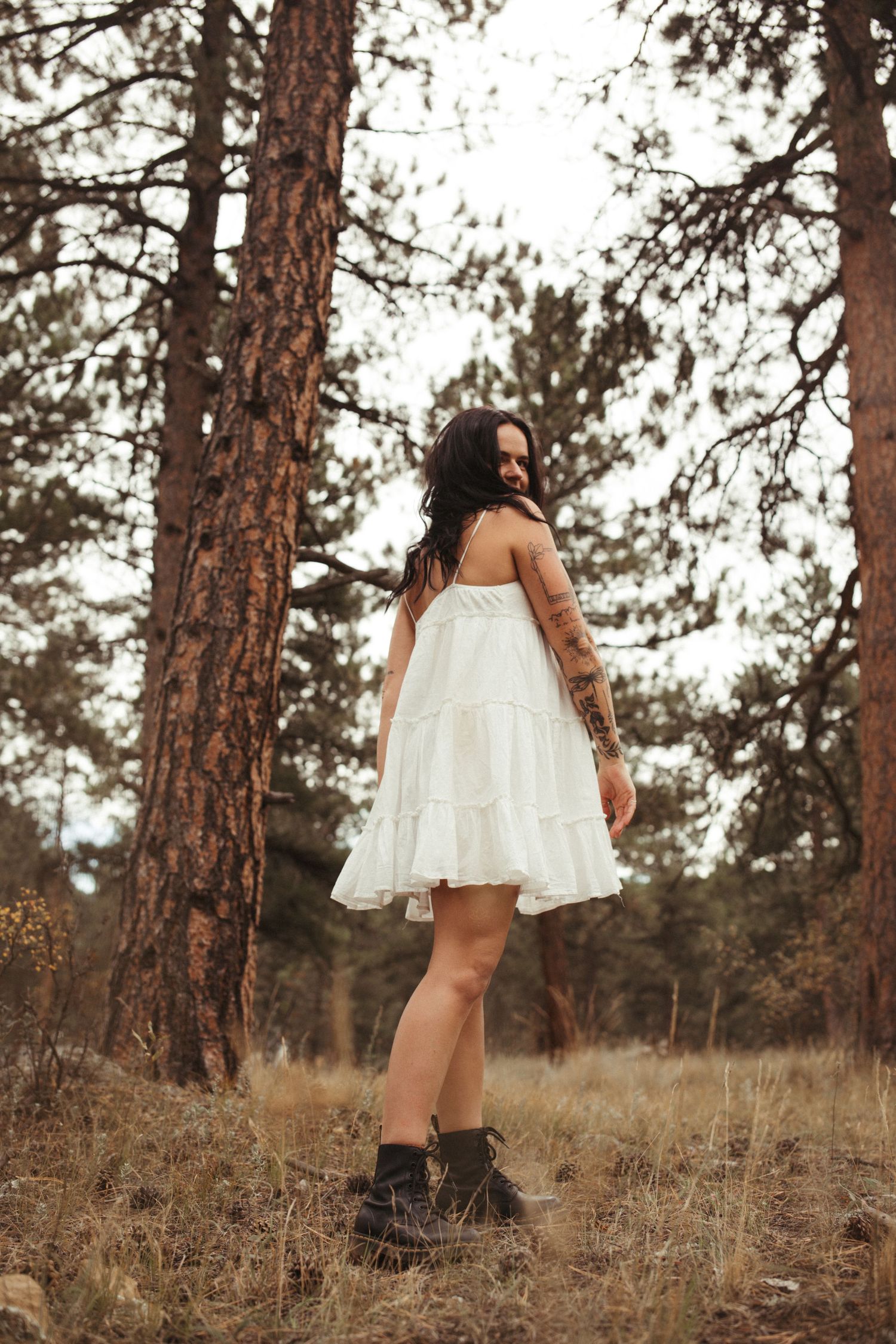 A person in a white dress poses among tall pine trees in a forest setting.