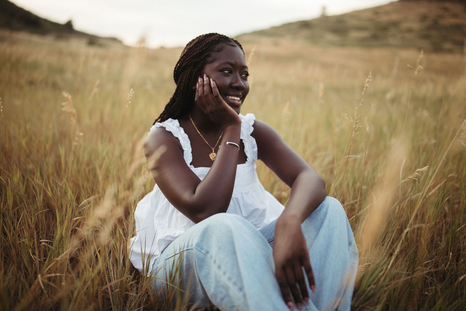 Someone in a white dress sits thoughtfully in a golden field at sunset, hand resting on their cheek.