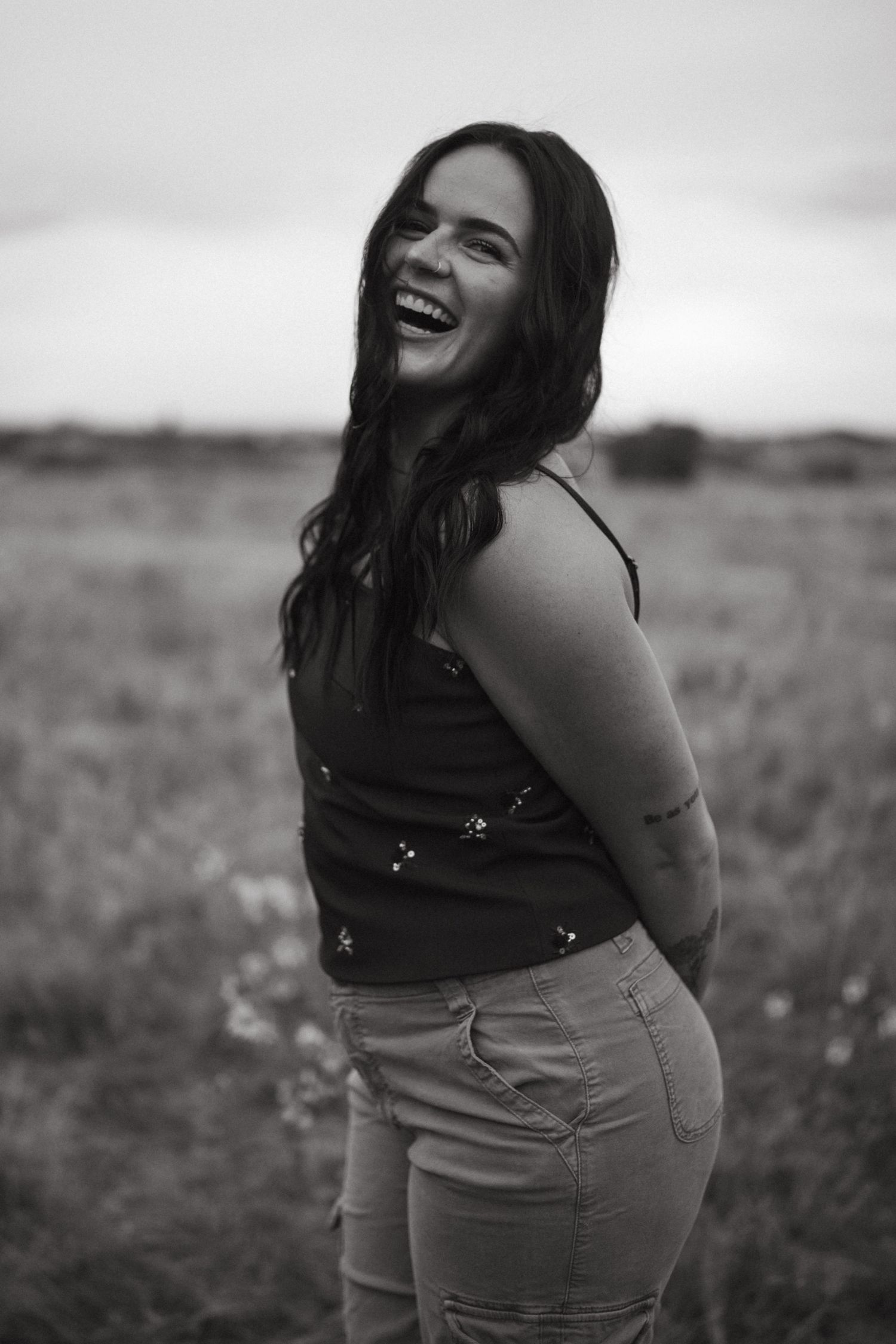A joyful person wearing a black top and khaki pants poses candidly in a field during a black and white photoshoot.