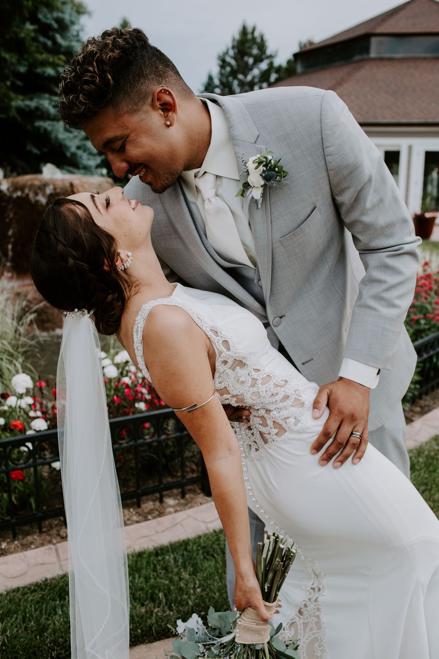 A romantic wedding photoshoot showing a couple in grey and white formal attire leaning against a white garden fence.
