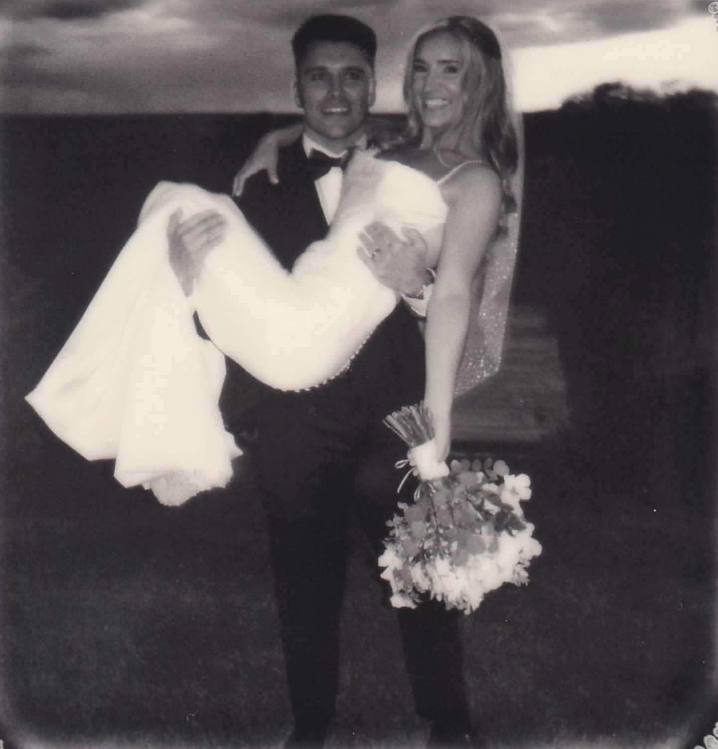 Groom in tuxedo carries bride in wedding dress holding bouquet, black and white outdoor wedding photo.