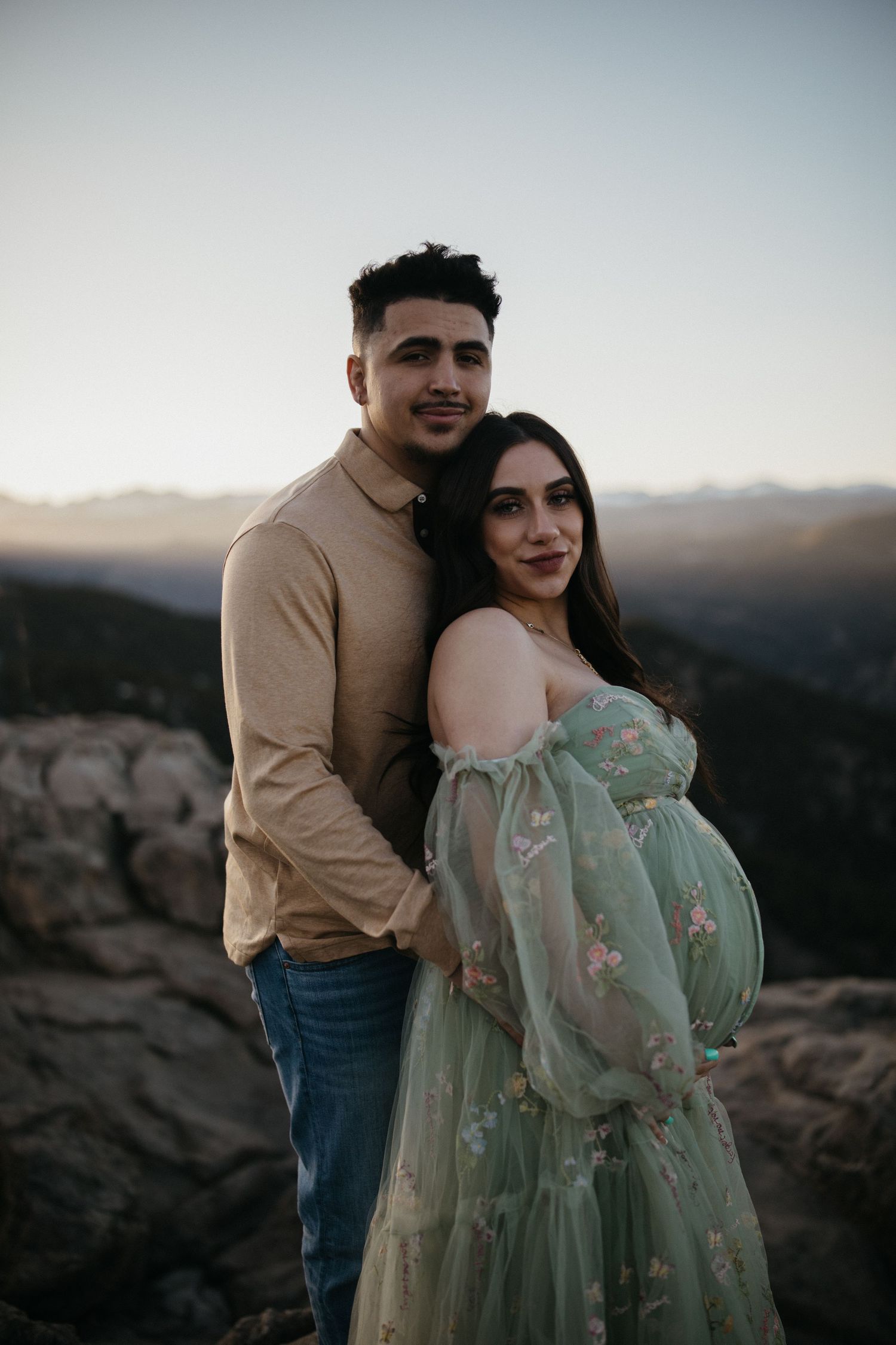 Sunset maternity photo with couple in formal wear standing on rocky mountain overlook.