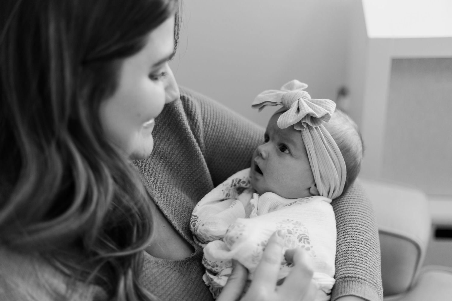 Mother in grey sweater holding and gazing lovingly at newborn baby wearing bow headband.