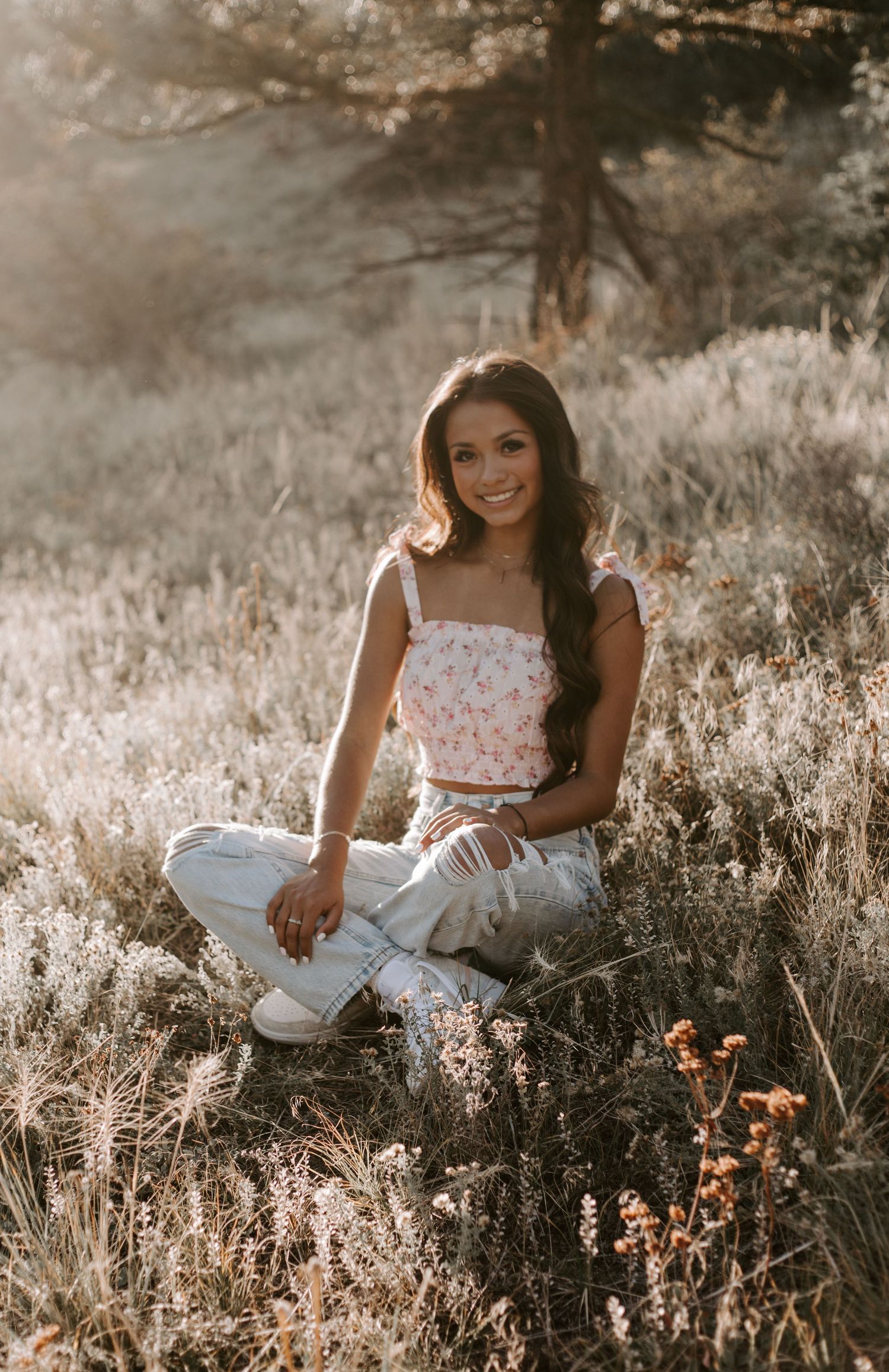 A summery portrait in a golden meadow shows someone in a floral crop top and white jeans during a dreamy sunset.