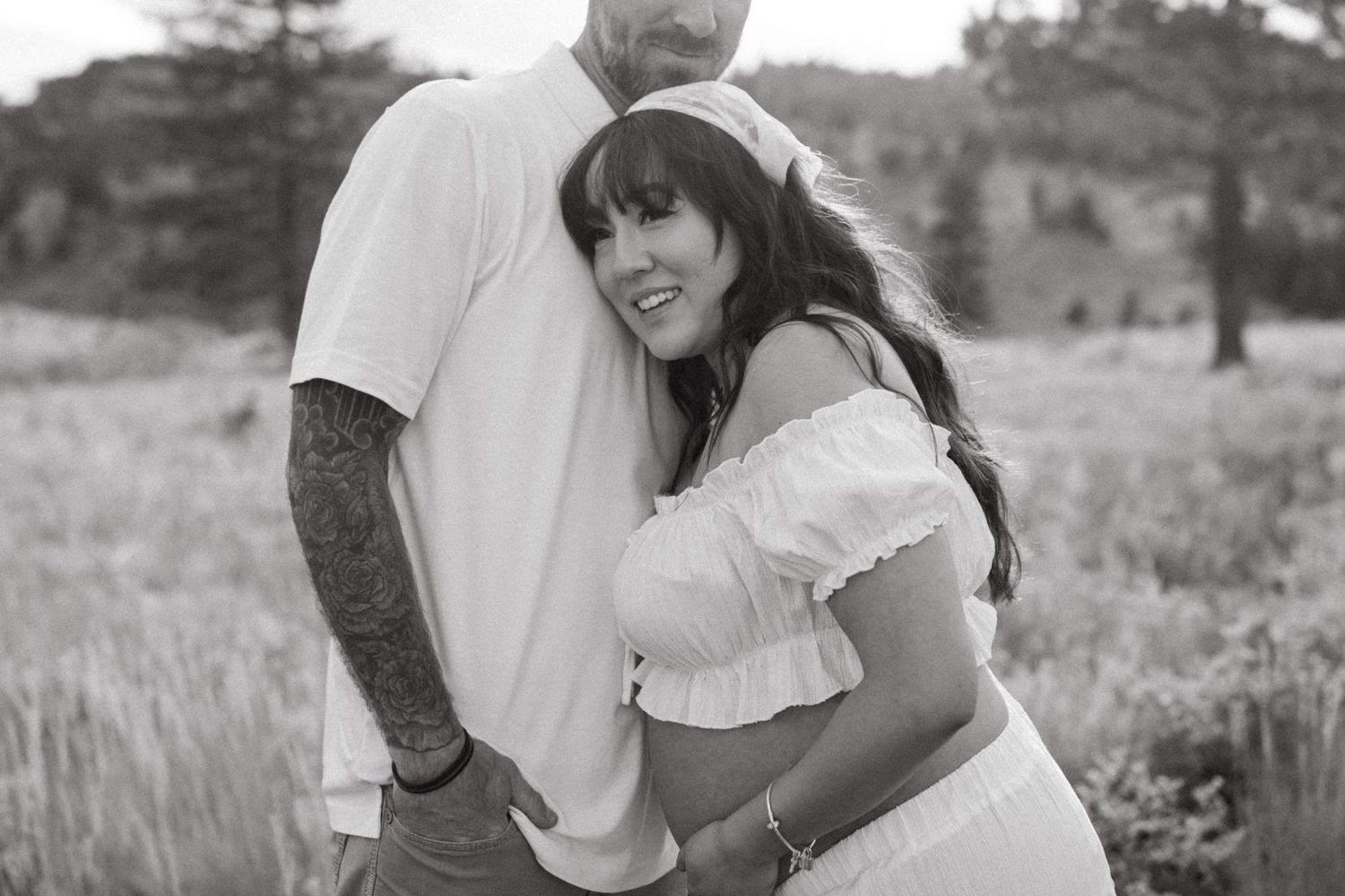 A couple embraces and laughs together in a sunlit field with tall grass and trees in the background.