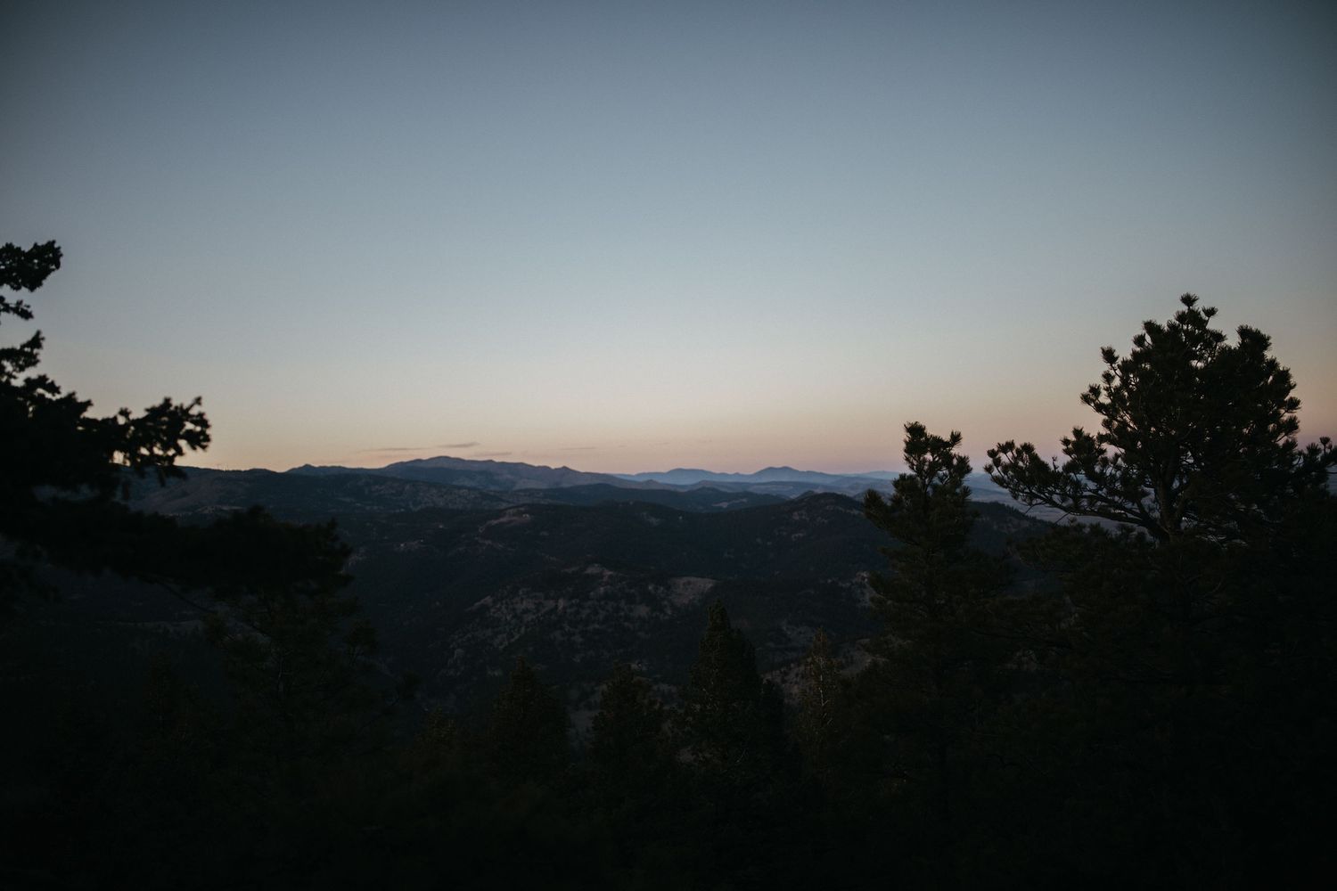 Mountain landscape silhouetted against a dusky sky with evergreen trees in the foreground.